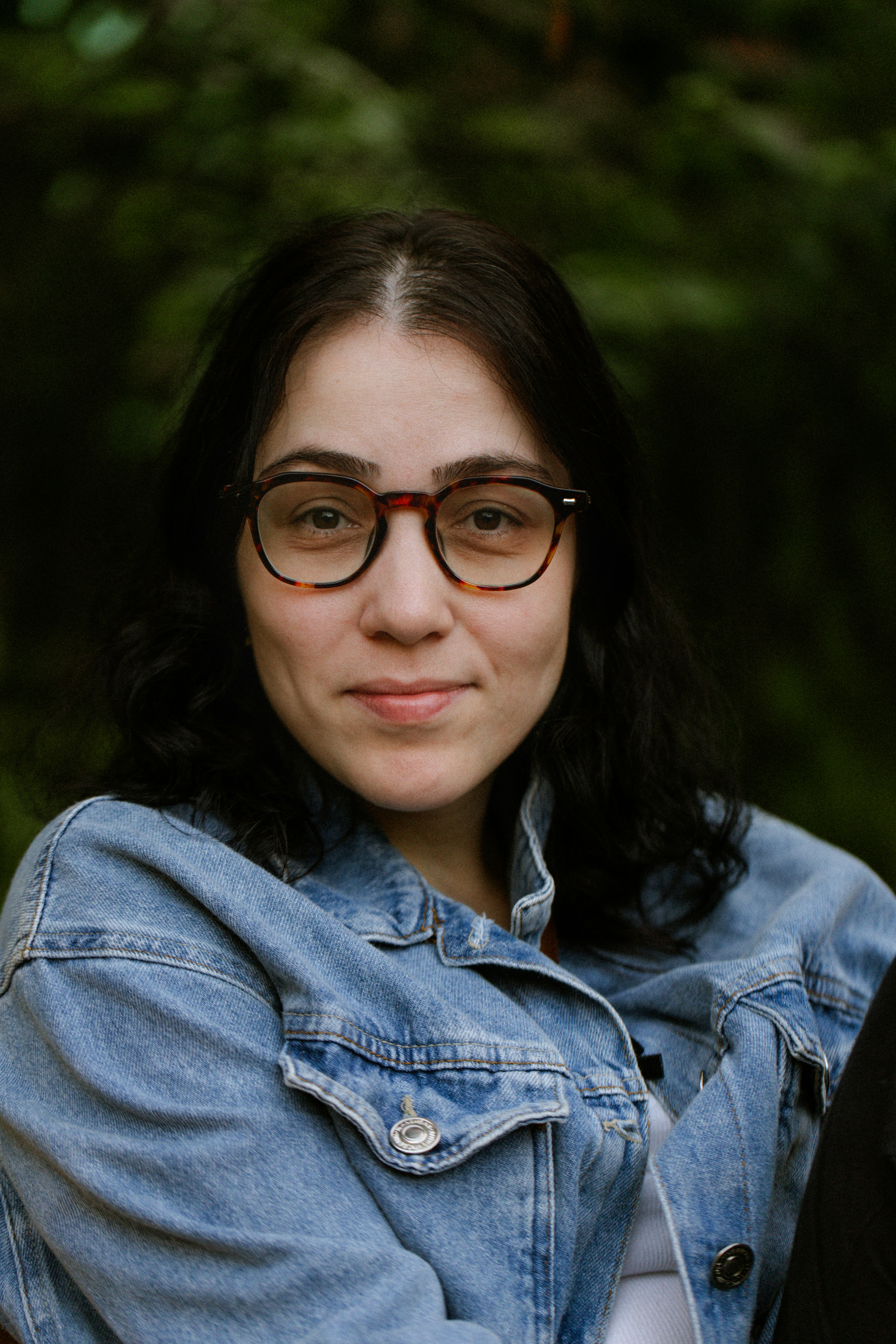 A woman with dark hair wearing tortoiseshell glasses and a denim jacket.