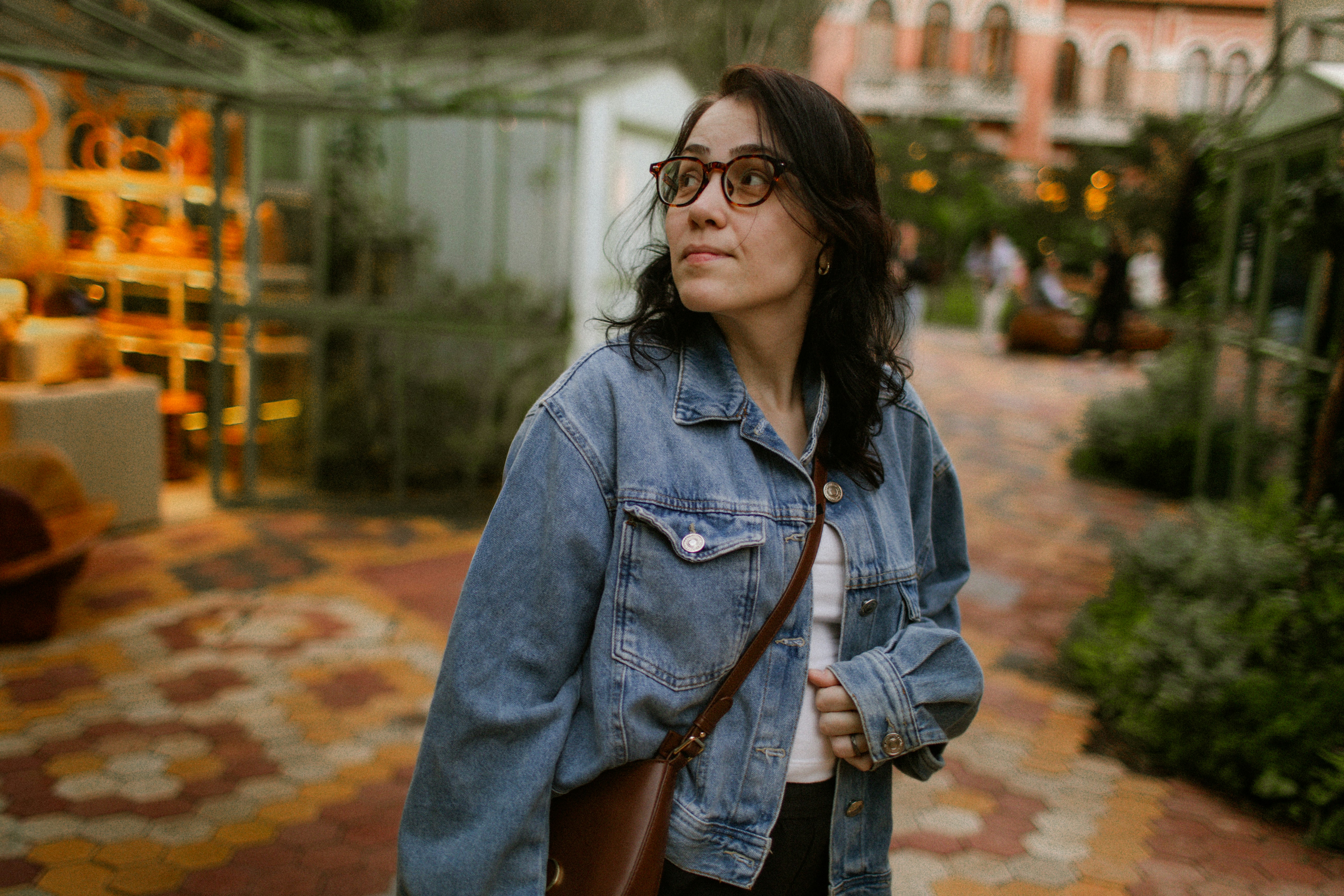 A woman in a denim jacket and glasses looking away in a garden.