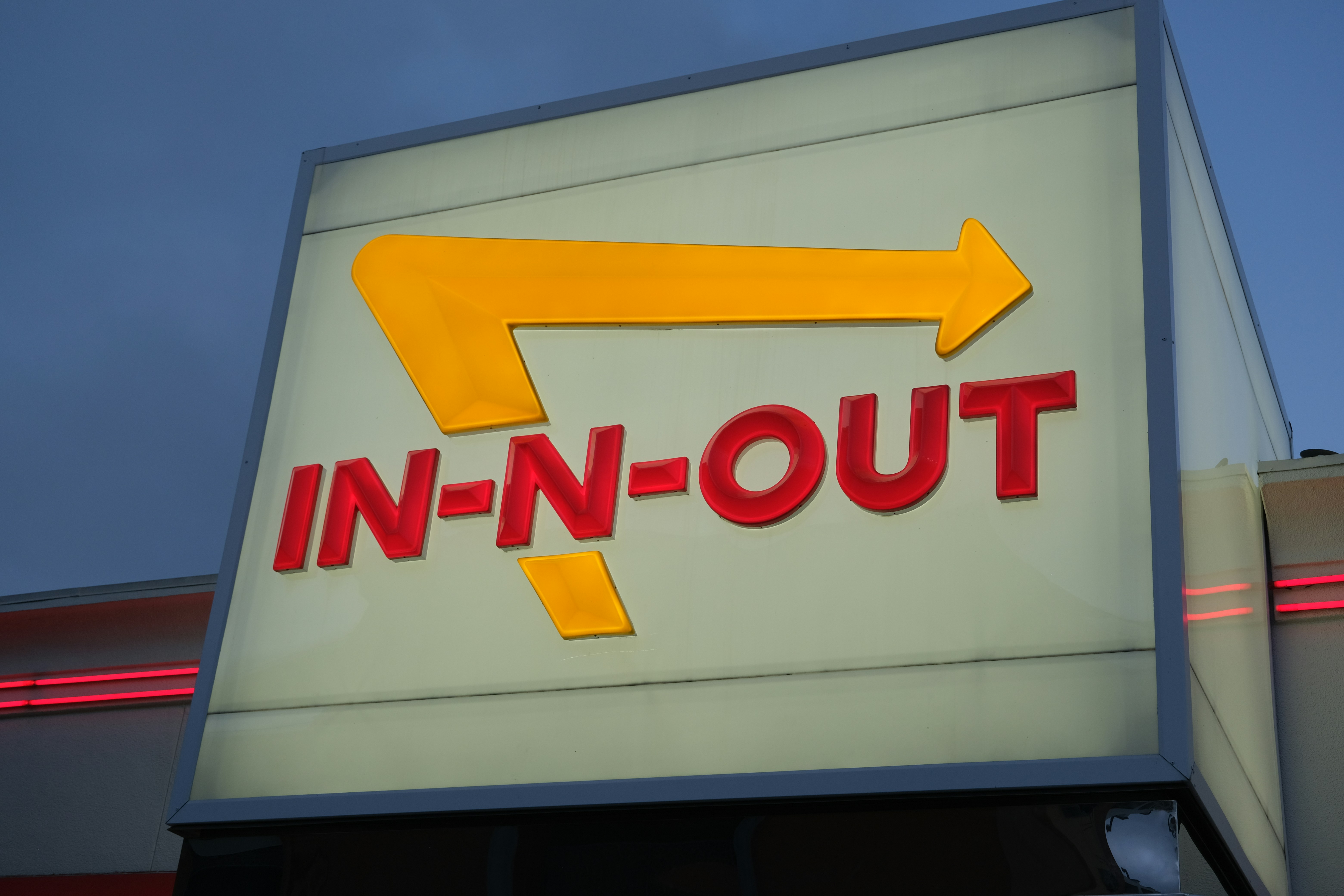 An illuminated In-N-Out Burger sign with a yellow arrow against a blue sky