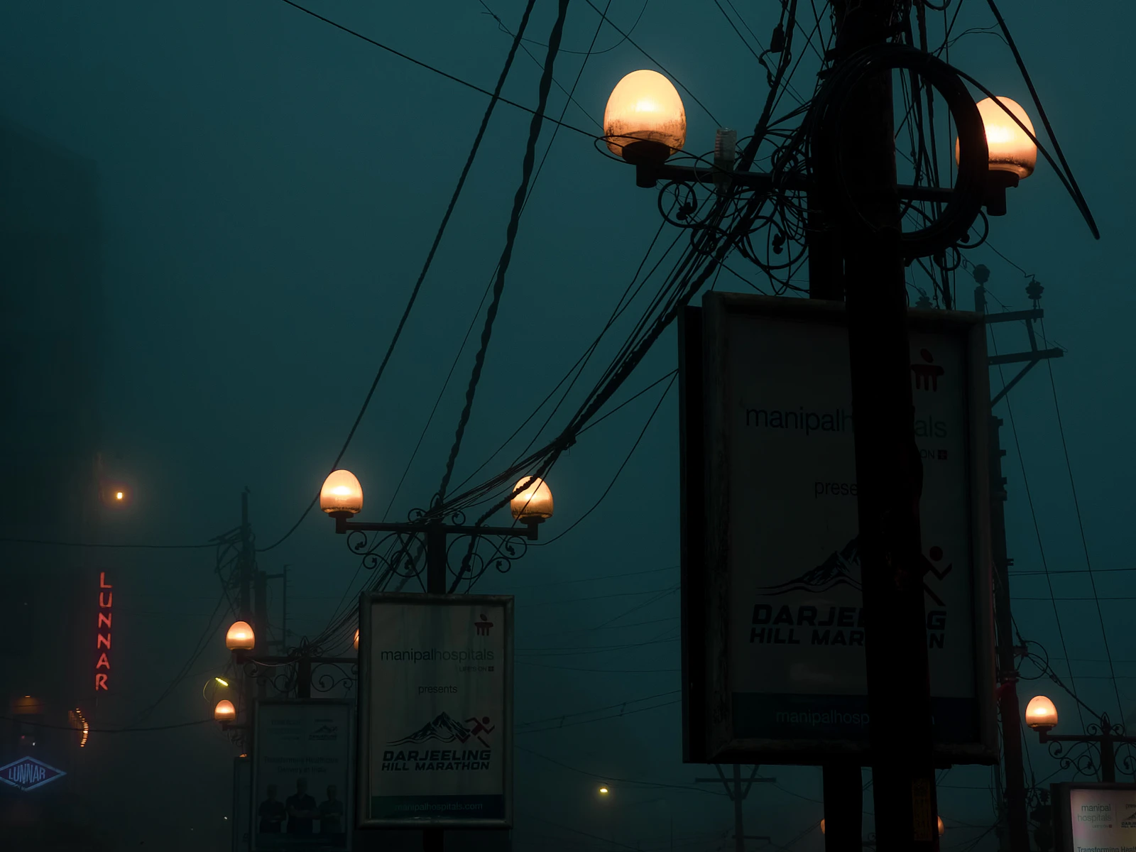 Street lamps glowing through thick fog above the signs of a Darjeeling street at night