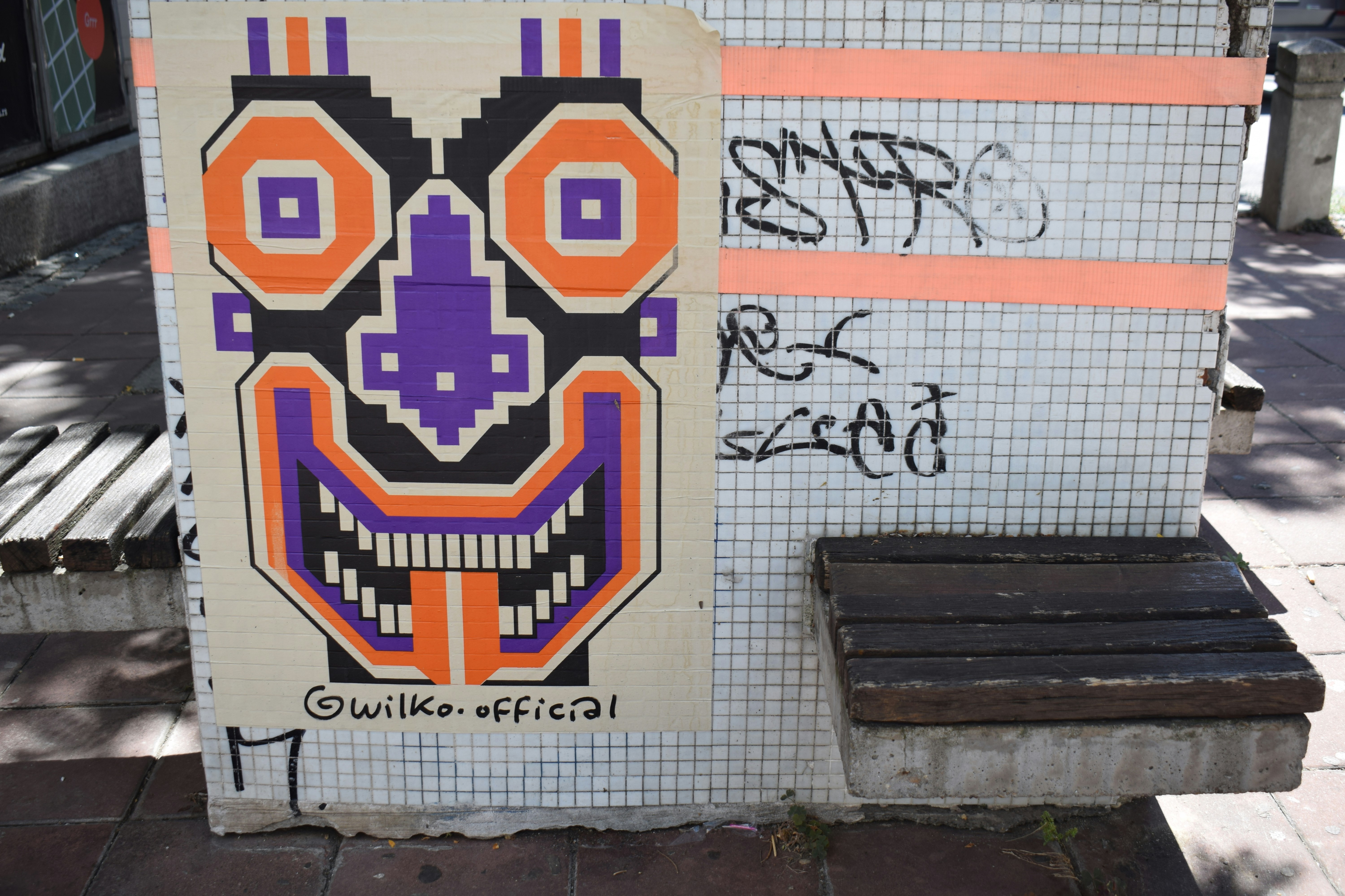 A pixelated orange and purple mask poster on a tiled wall with graffiti