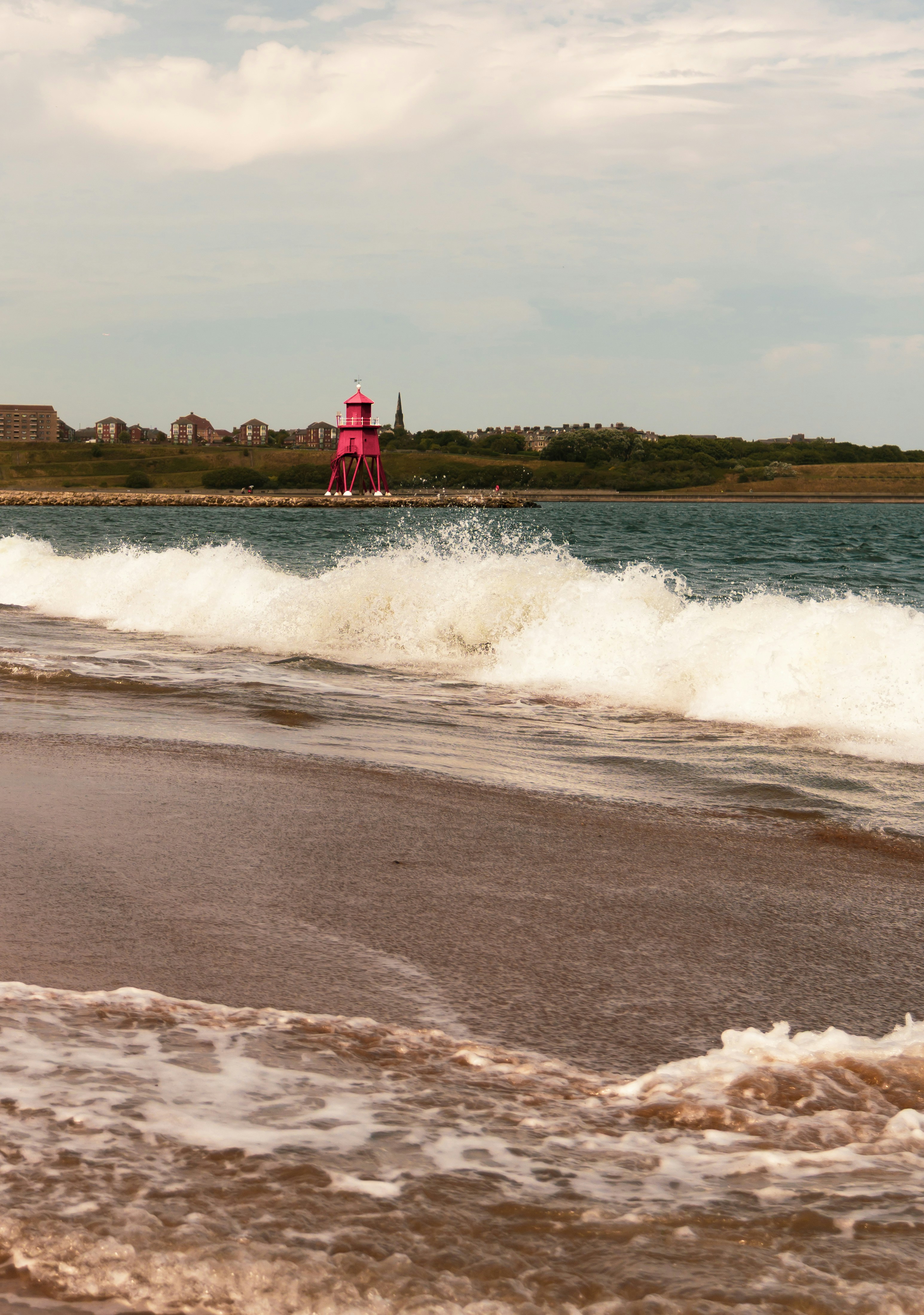 A bright pink lighthouse on a stone pier behind crashing ocean waves
