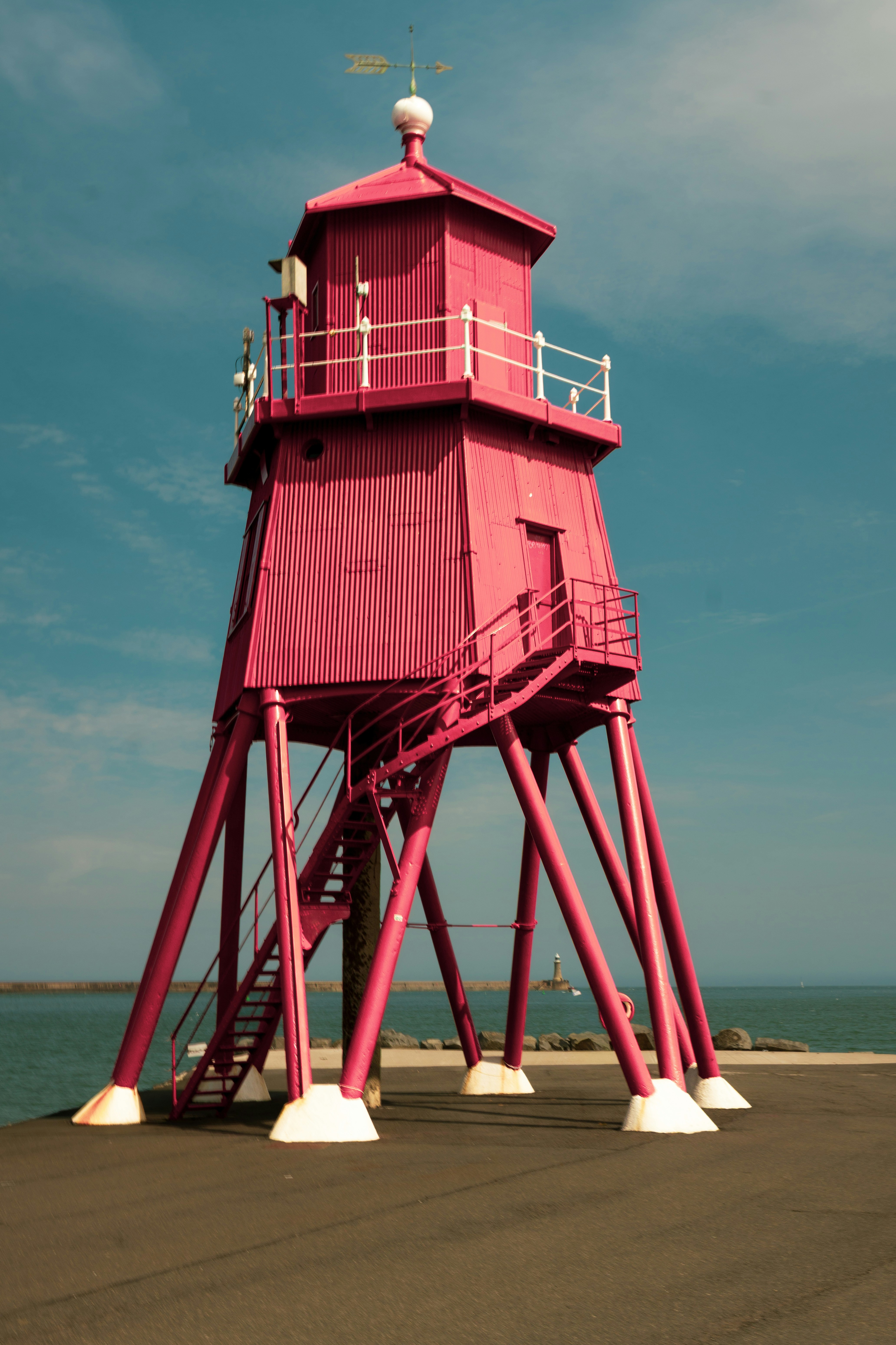 A bright pink lighthouse on stilts above a concrete pier by the sea