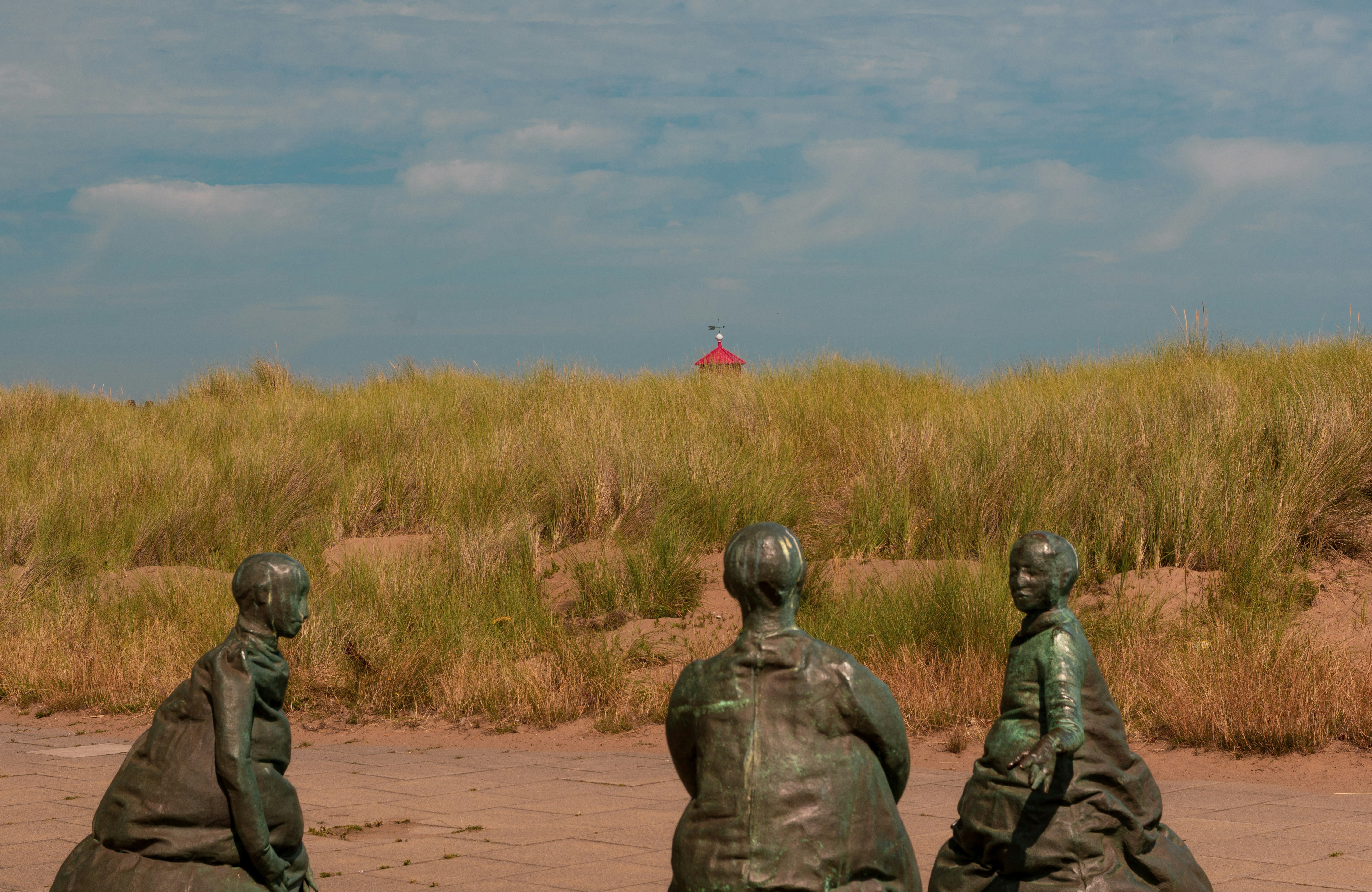 Three bronze statues of people facing a grassy dune with a red roof