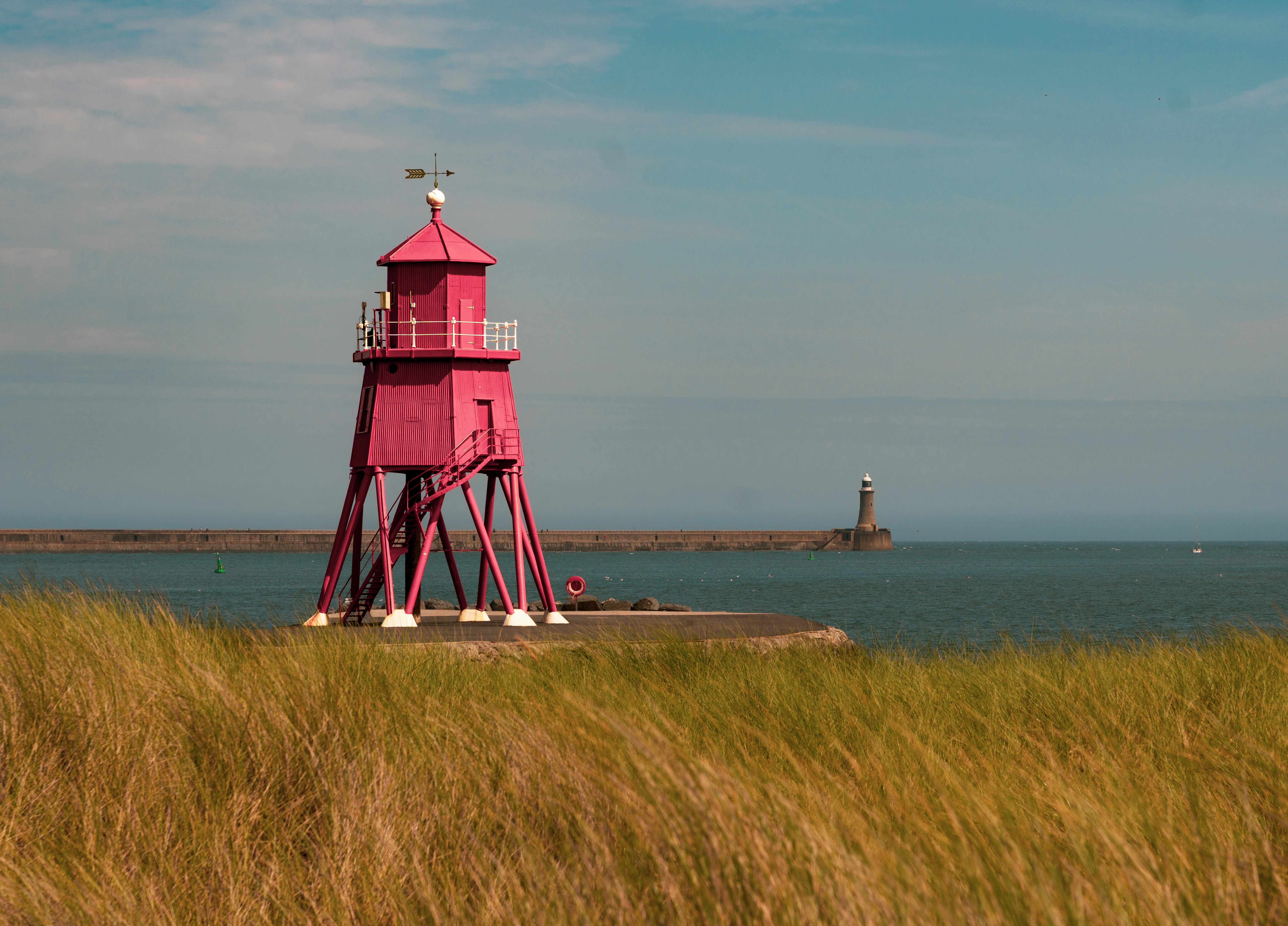 A bright pink lighthouse on stilts above a grassy dune near the sea