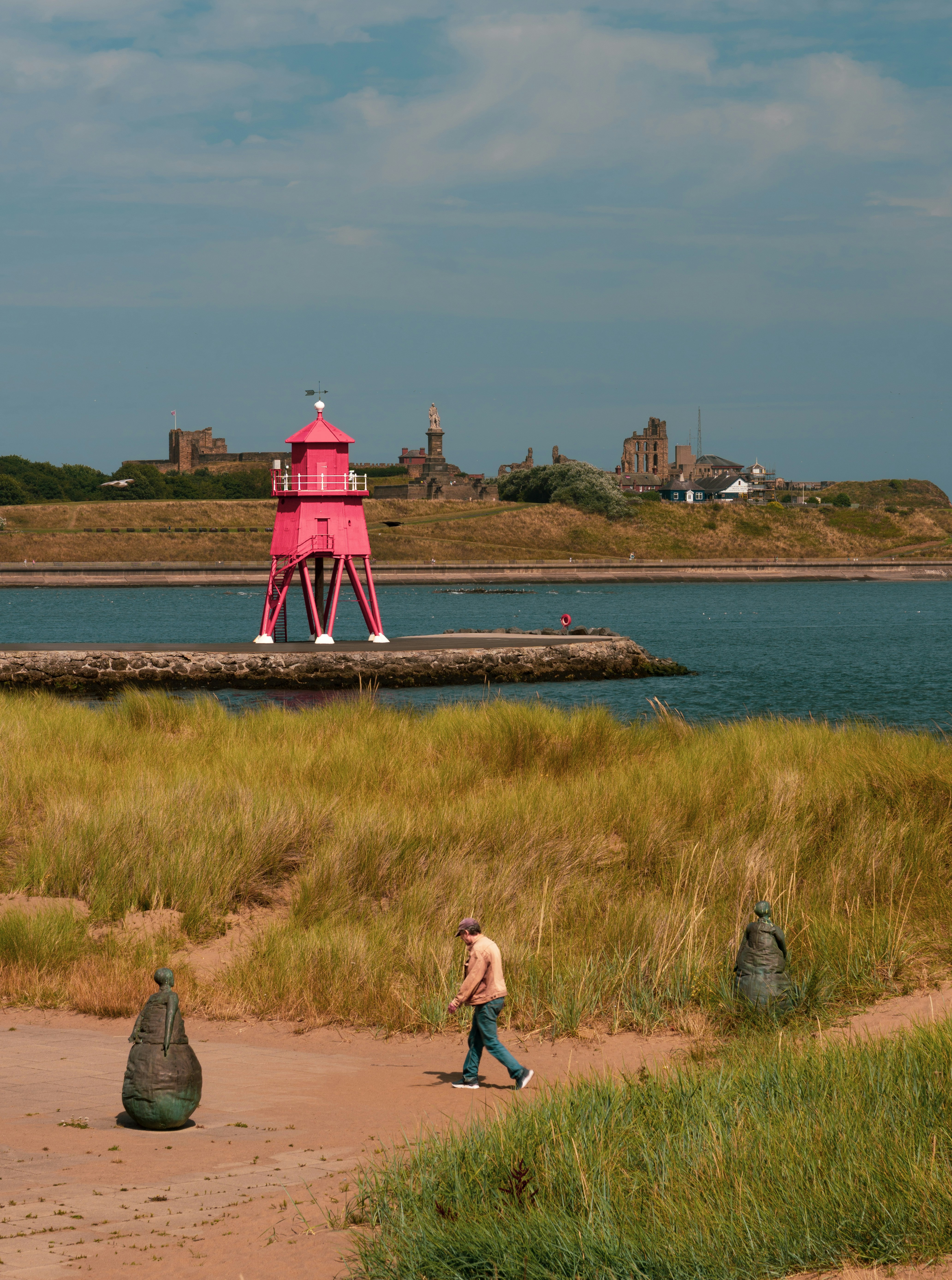 A person walking past bronze sculptures on a sandy path near a pink lighthouse