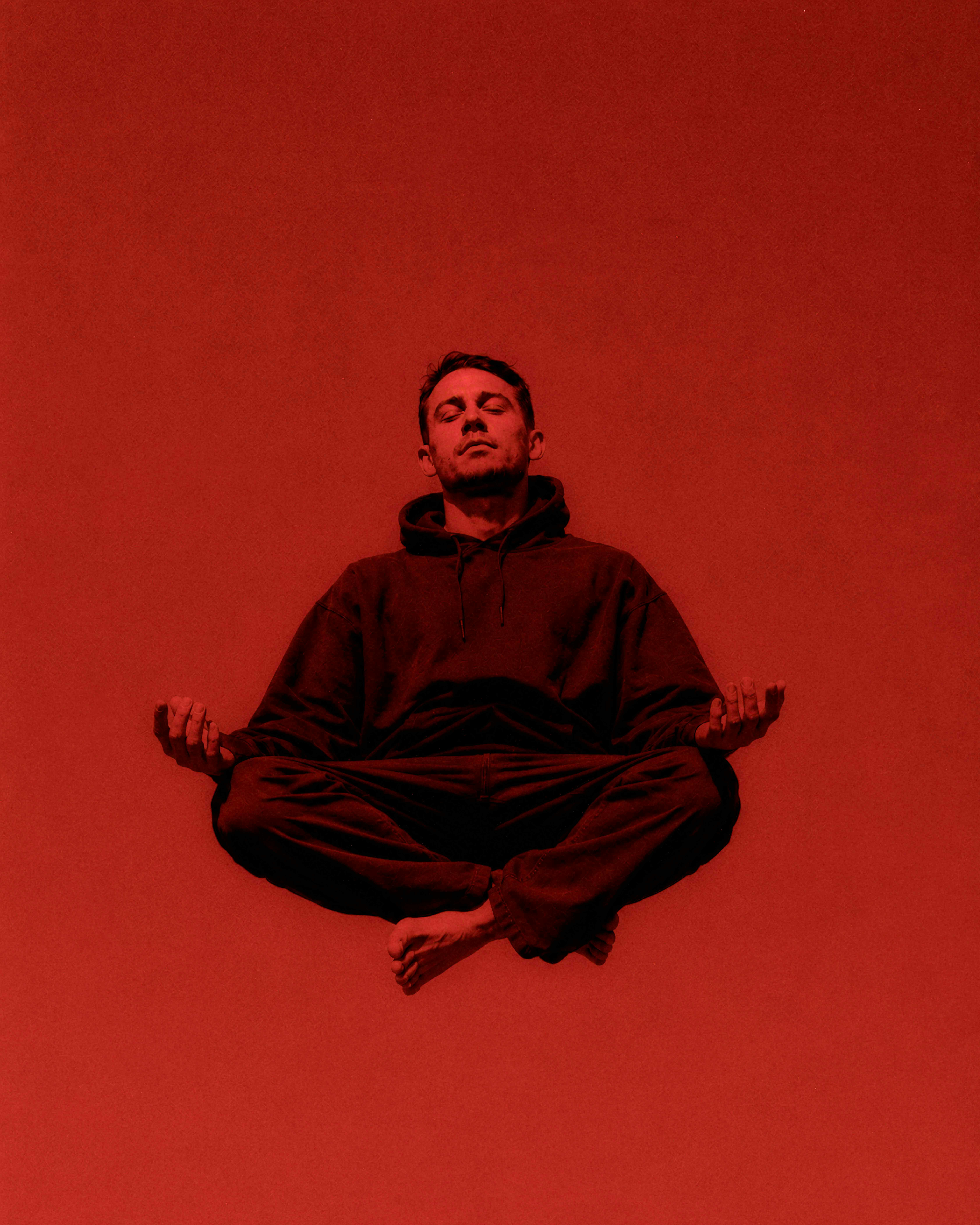 A man in a dark hoodie meditating in a cross-legged position against red light