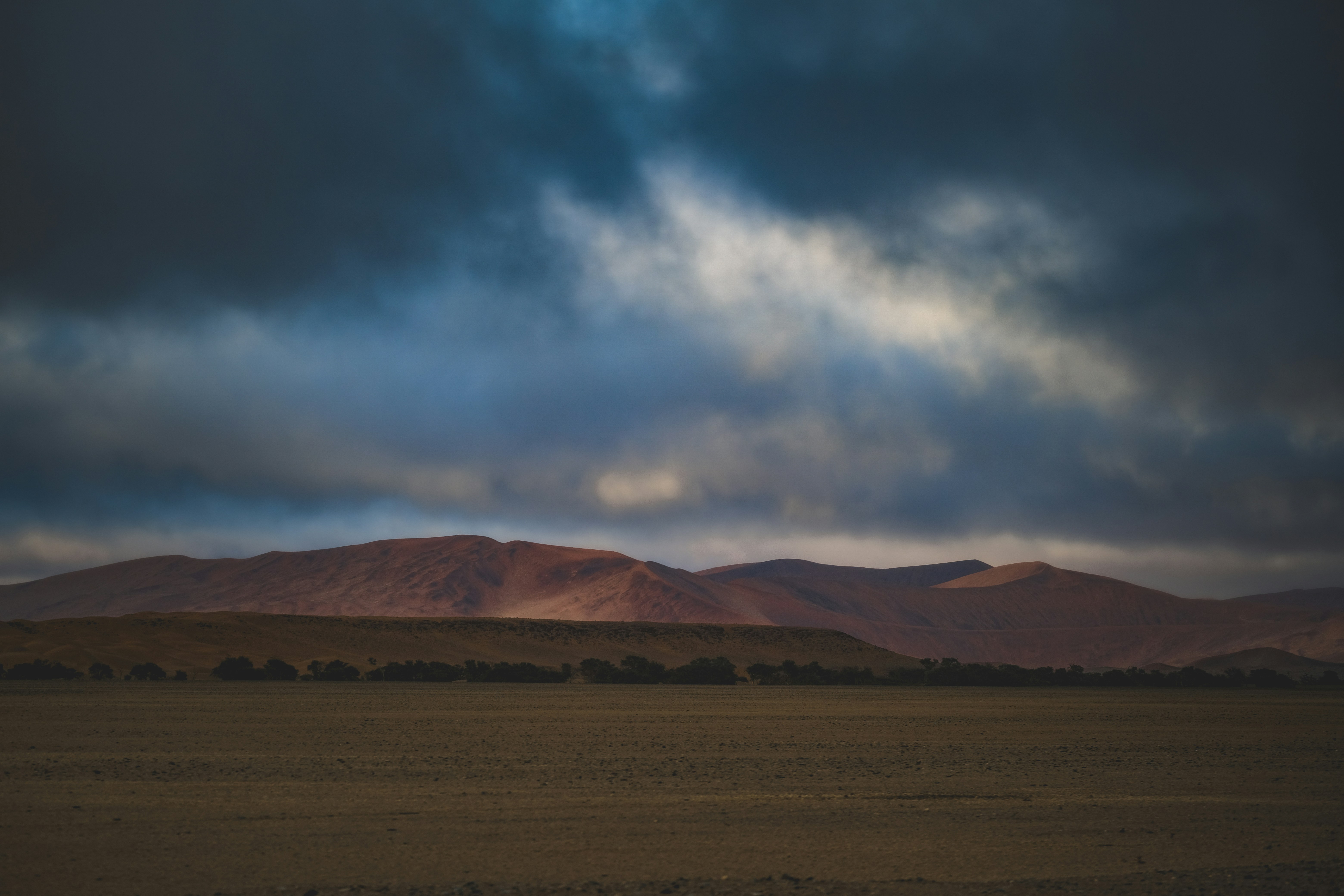 Reddish brown mountains rising above a flat desert plain under a dark cloudy sky