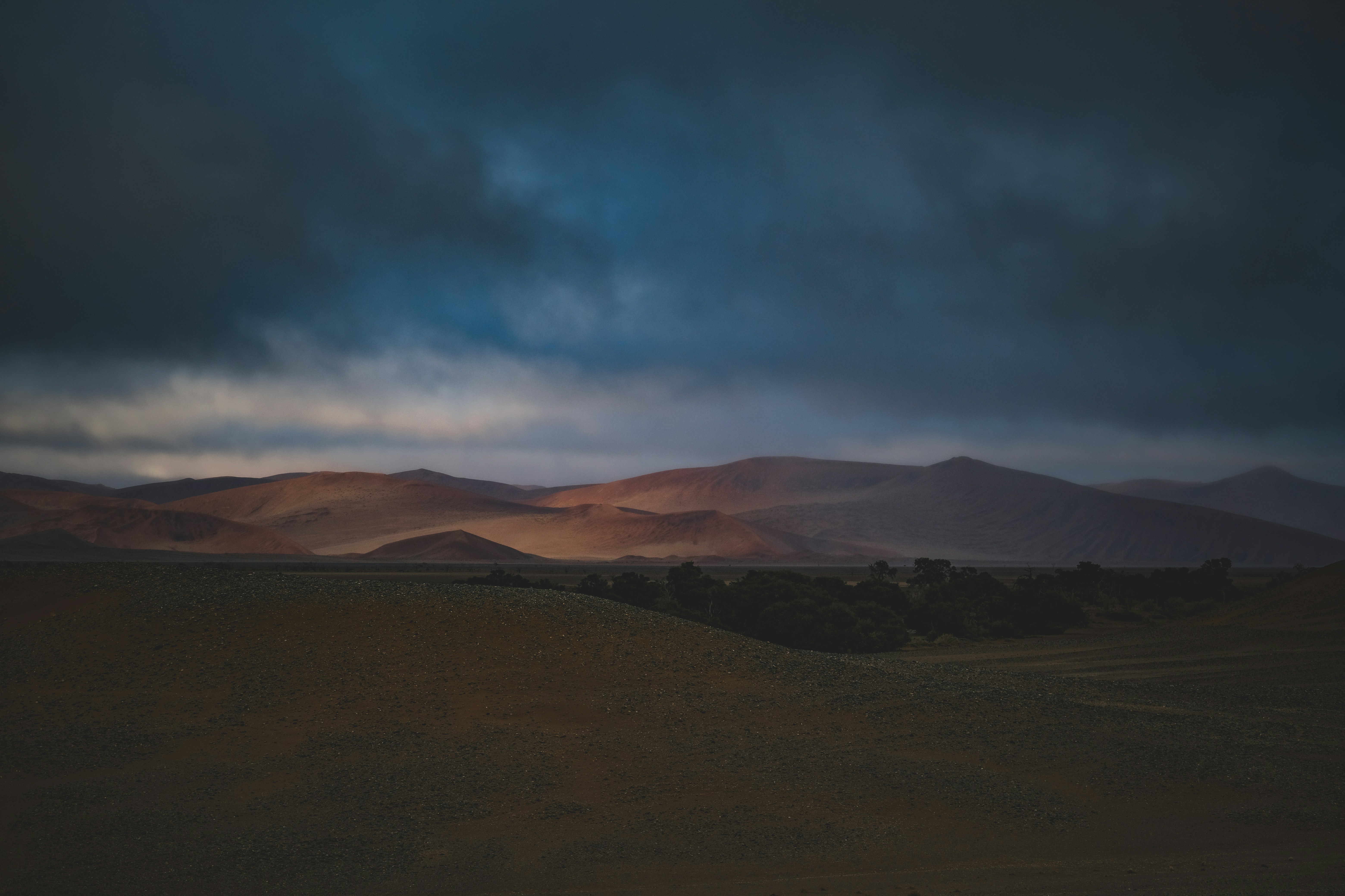Reddish desert mountains under a dark, heavy cloud layer at low light
