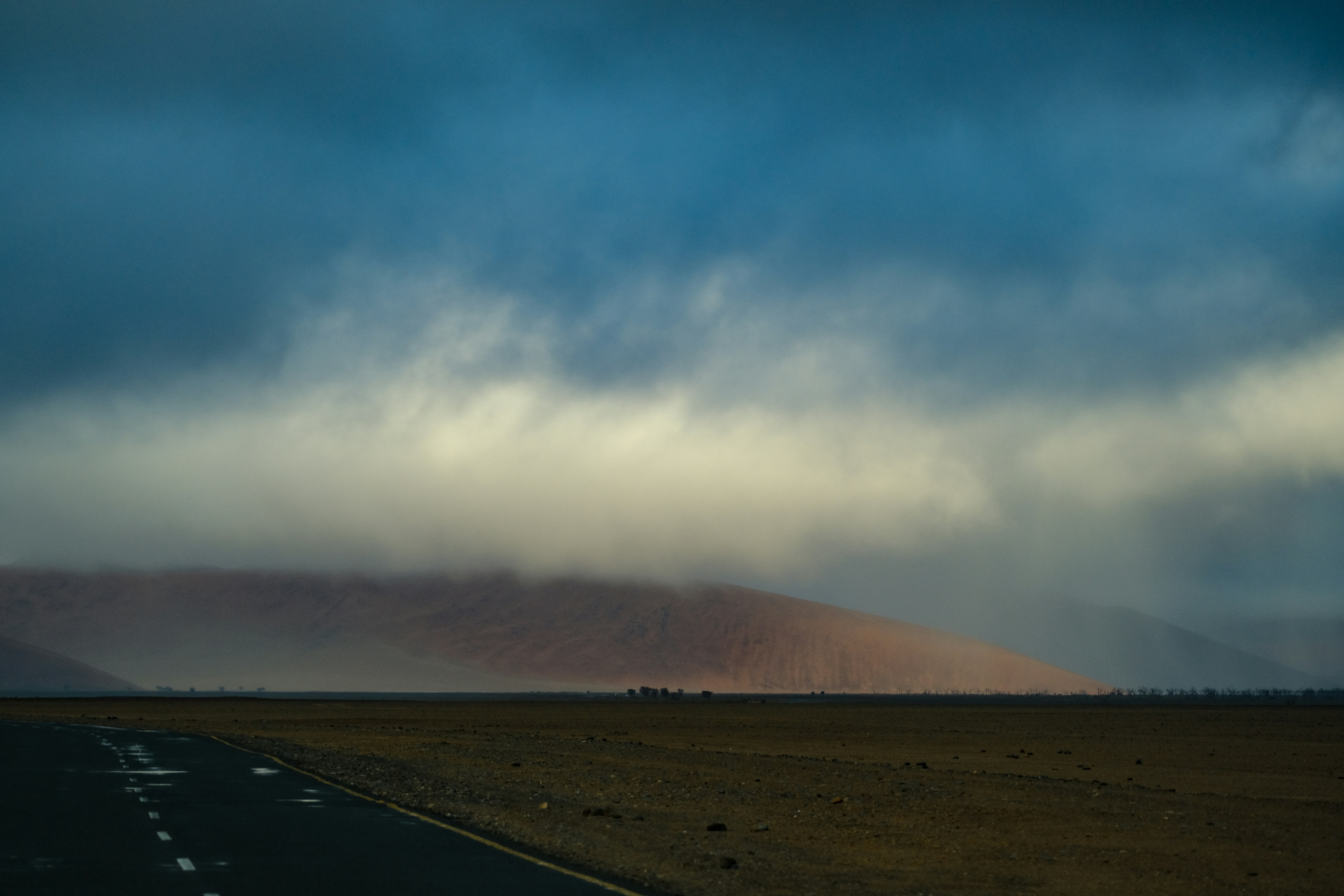 A paved road leading toward a large desert dune under heavy storm clouds