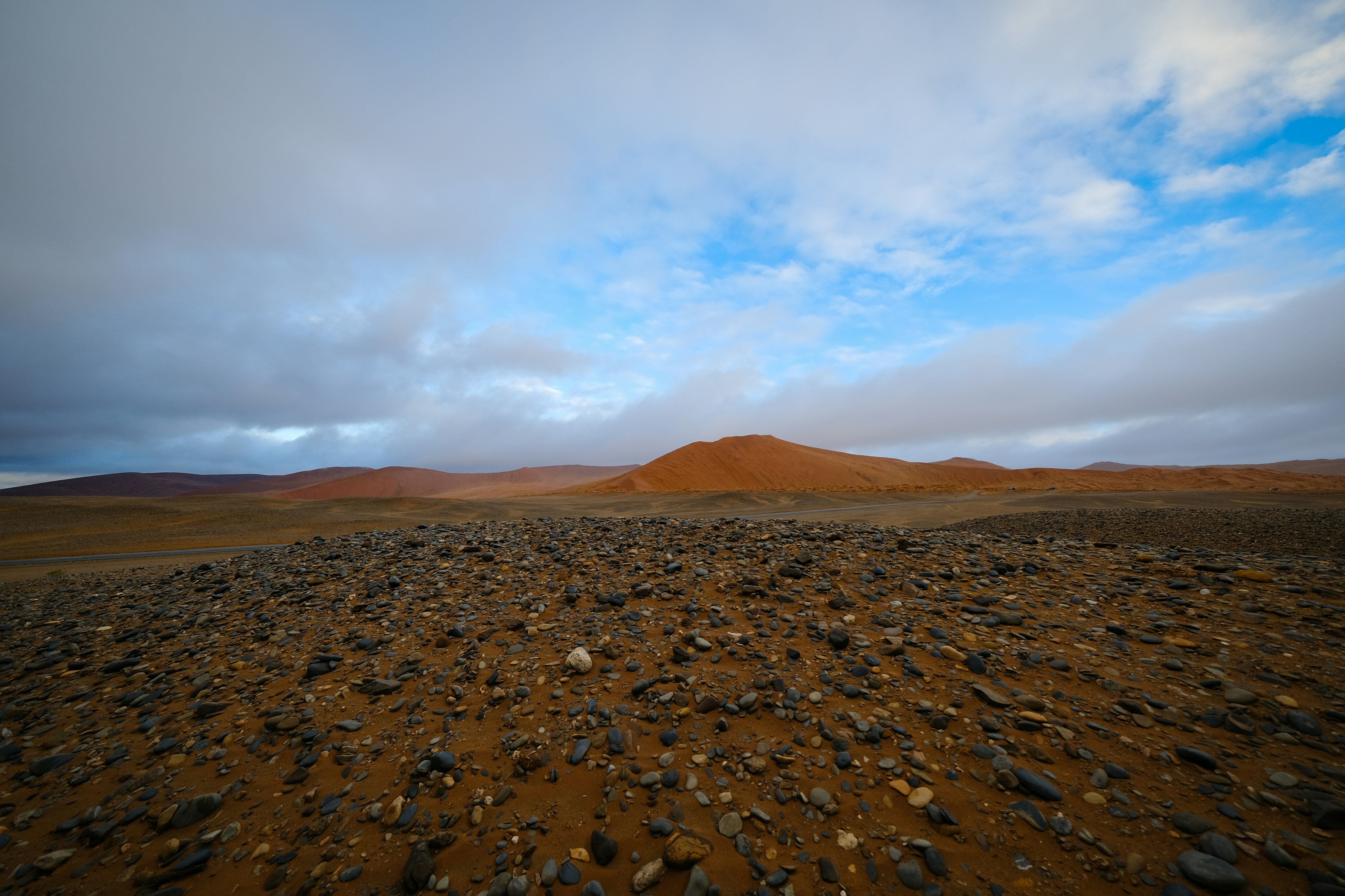 A rocky desert landscape with orange dunes under a cloudy blue sky