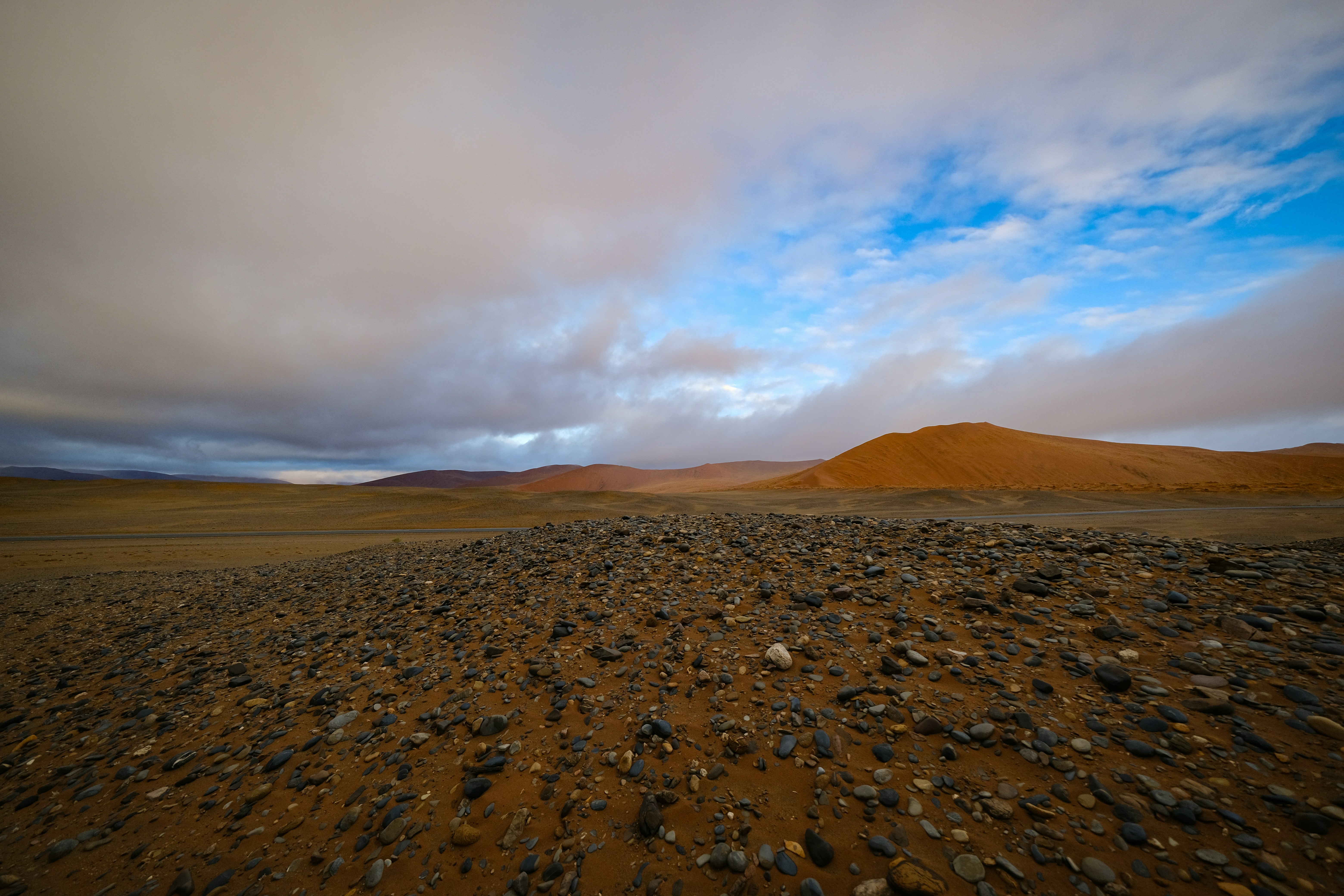 A rocky desert landscape with distant orange dunes under a cloudy blue sky