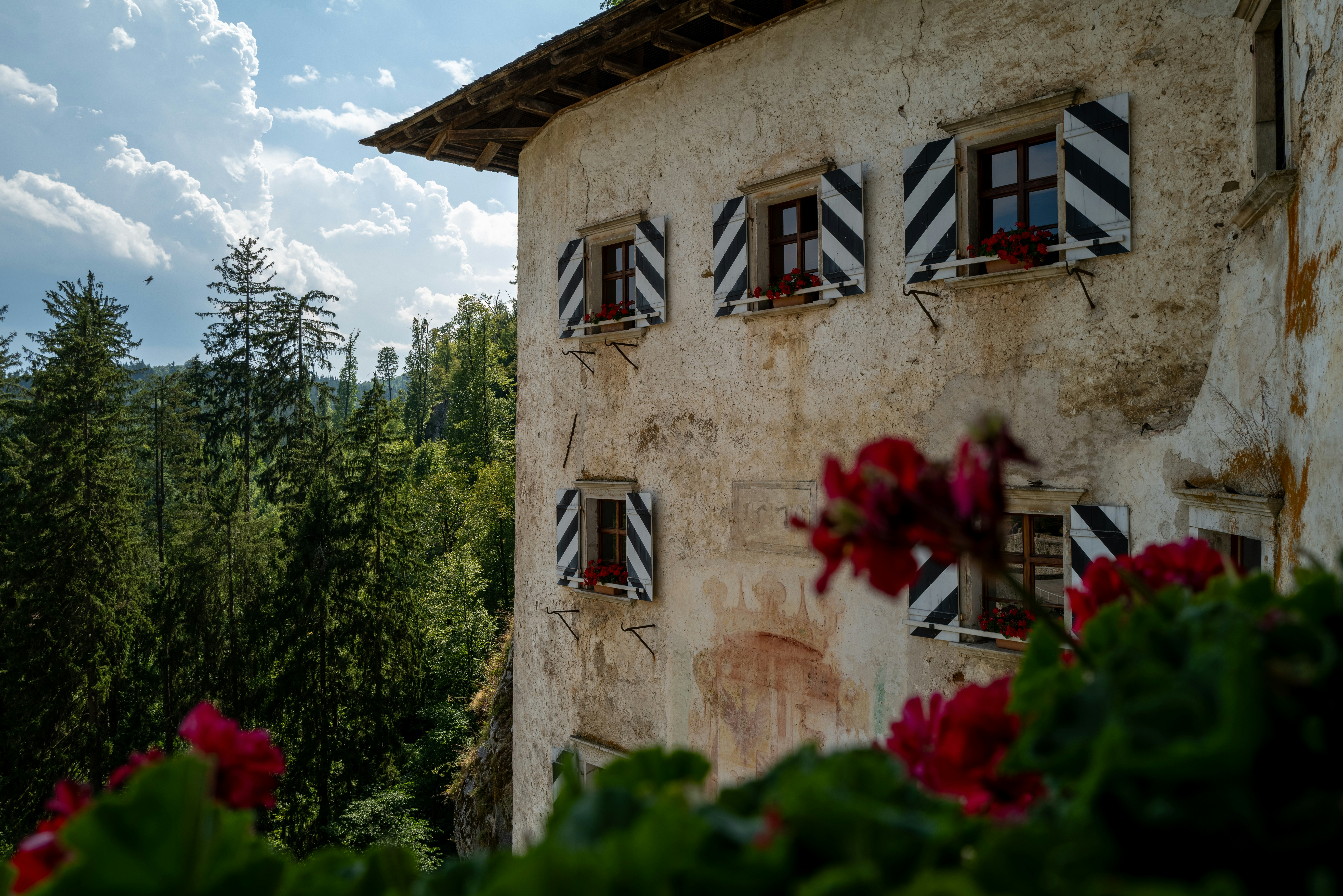 Ein altes Steingebäude mit gestreiften Fensterläden und roten Blumen in einem Wald