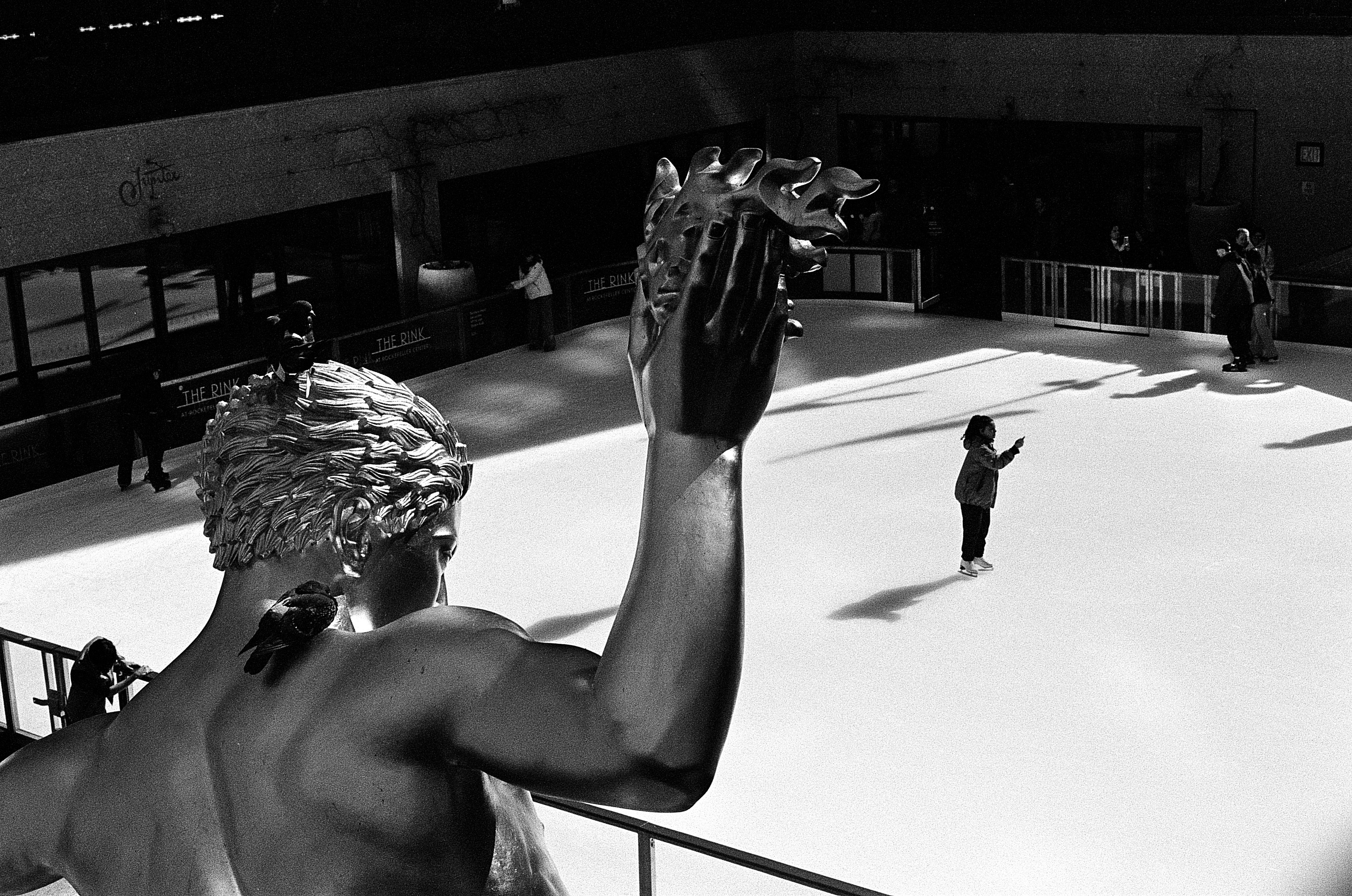 A bronze statue overlooking an ice rink with people skating in New York City