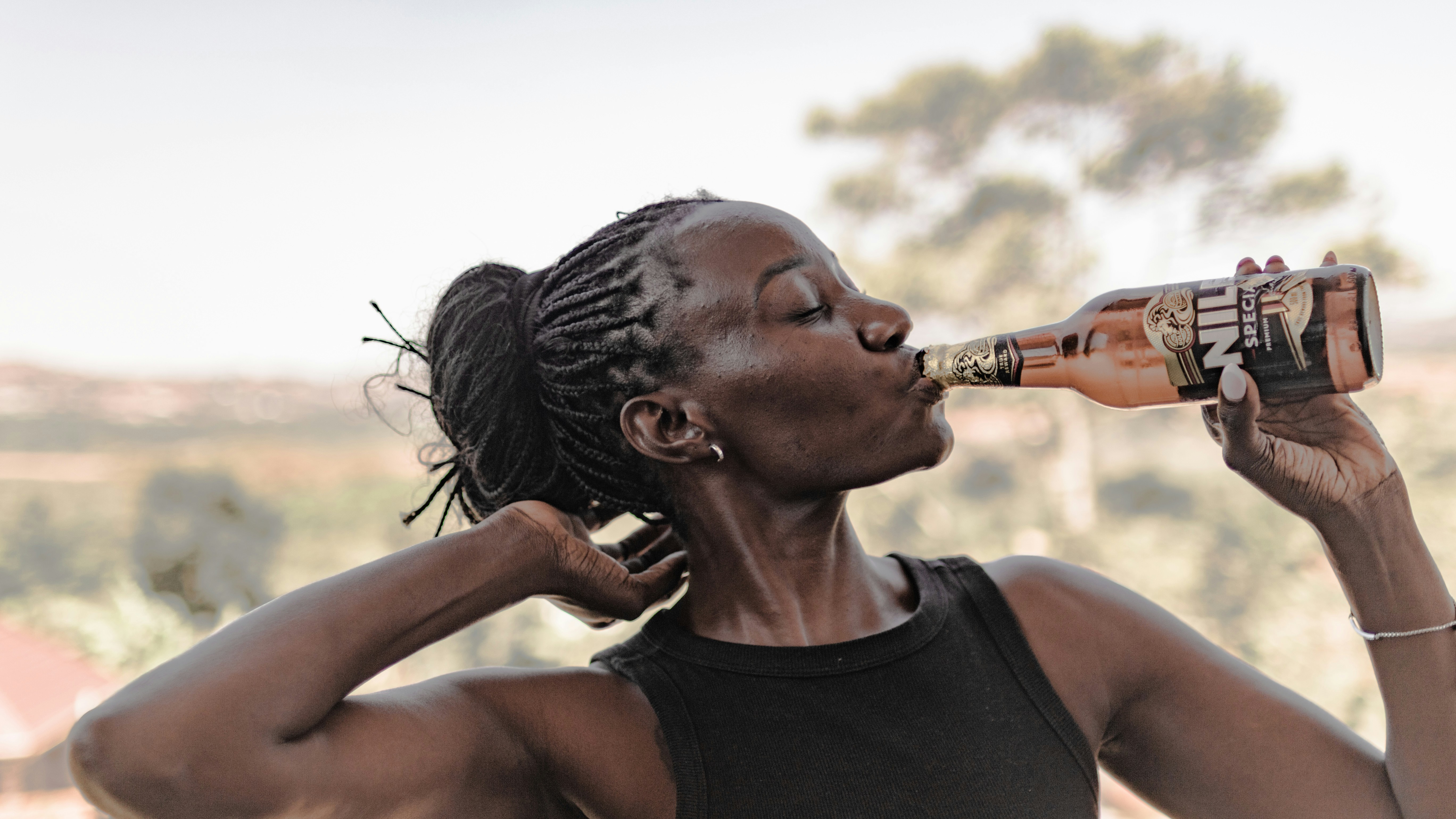 A woman with braided hair drinking from a brown glass beer bottle outdoors