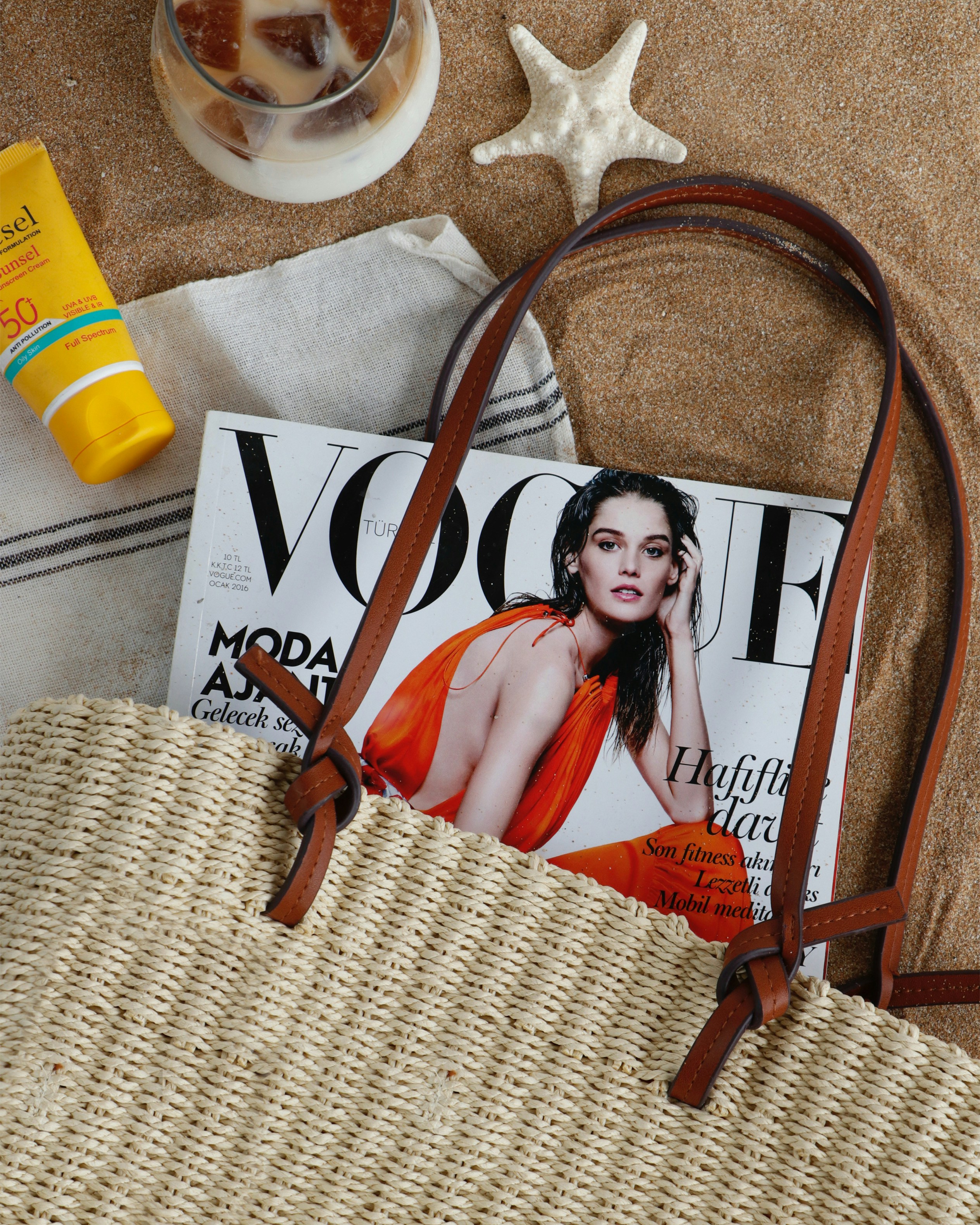 A straw tote bag, a fashion magazine, sunscreen, and a starfish on sand