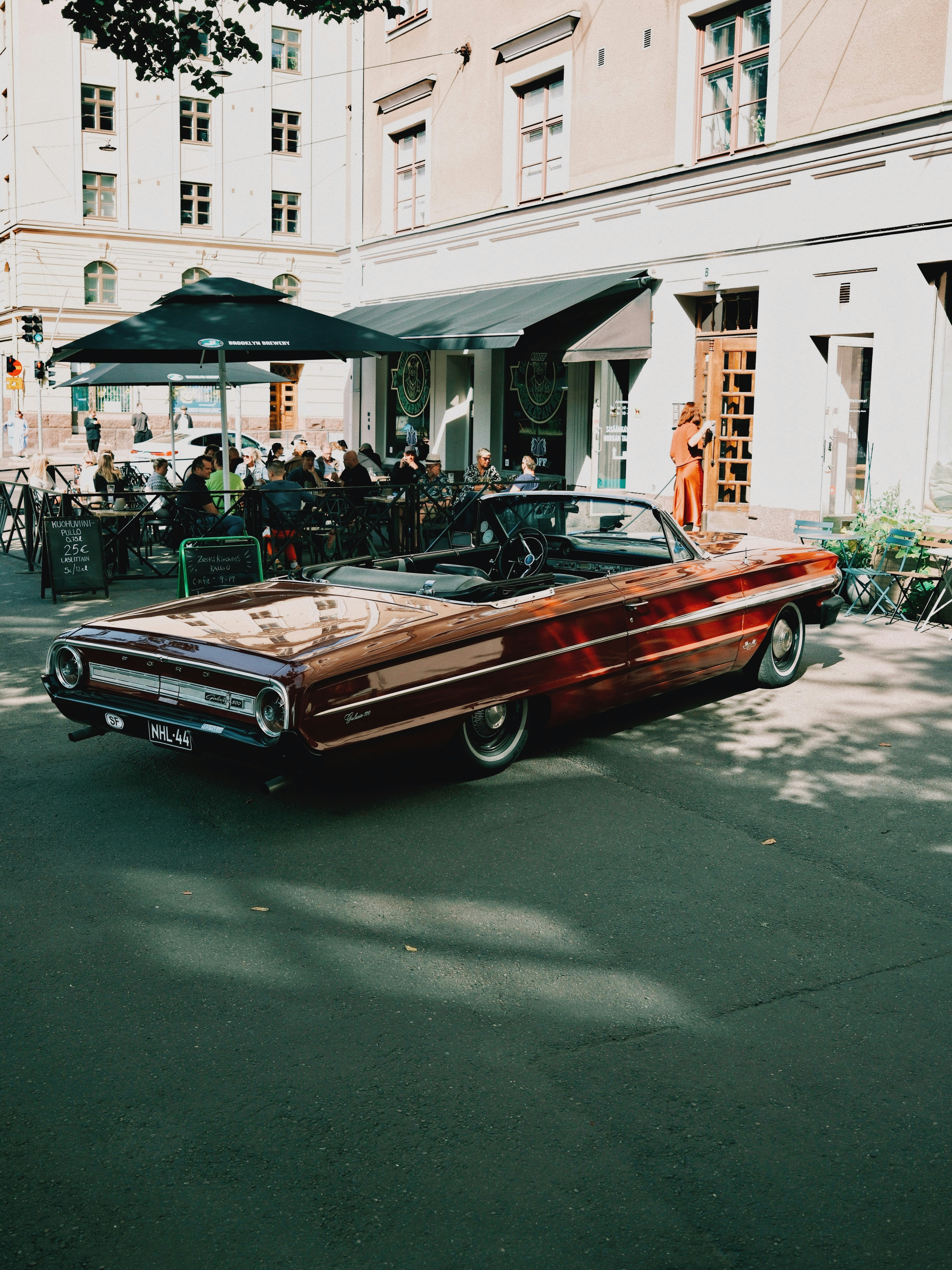 A vintage maroon Ford Galaxie convertible parked on a street near a cafe