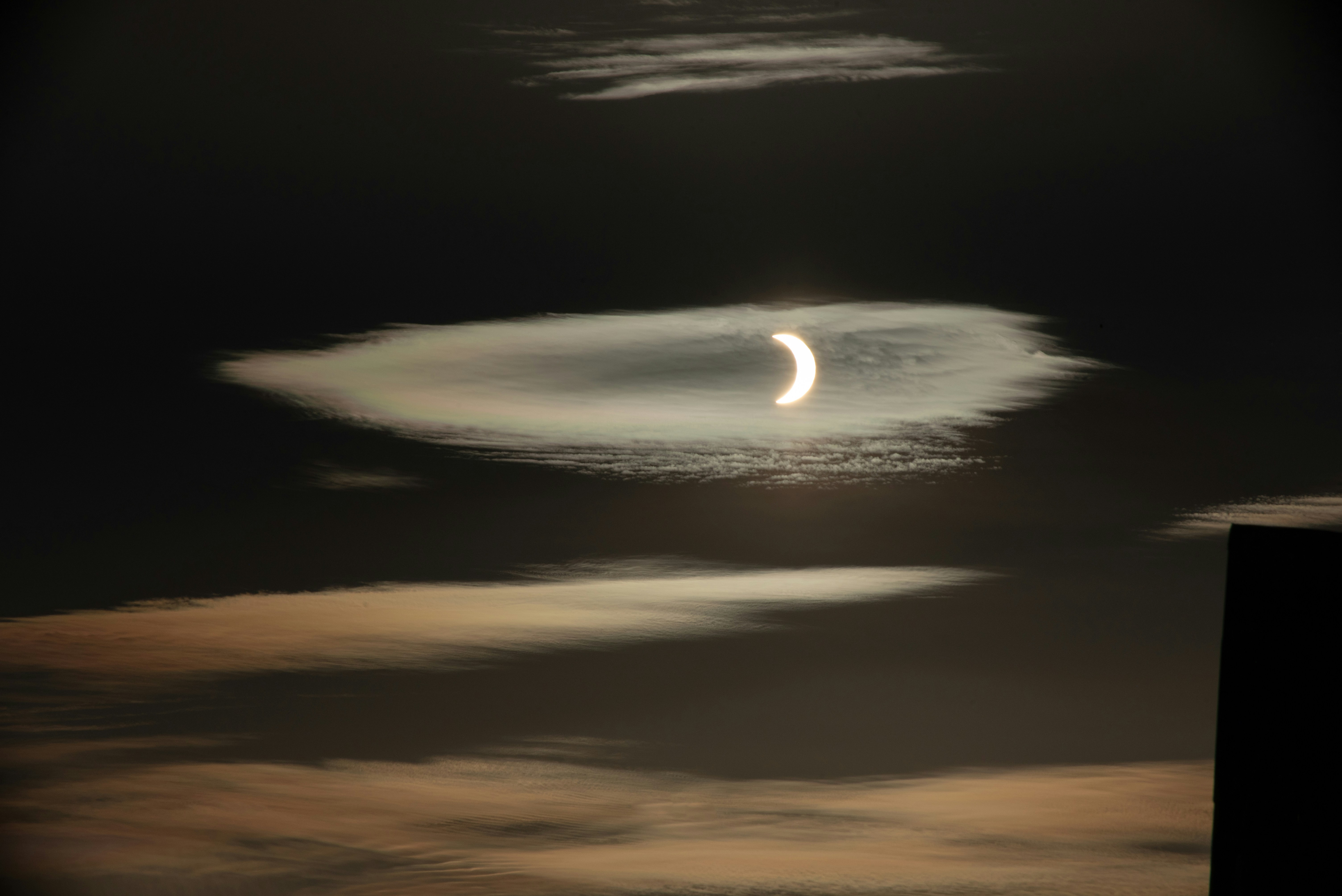 Un sole a mezzaluna parzialmente oscurato dalla luna dietro sottili nuvole iridescenti