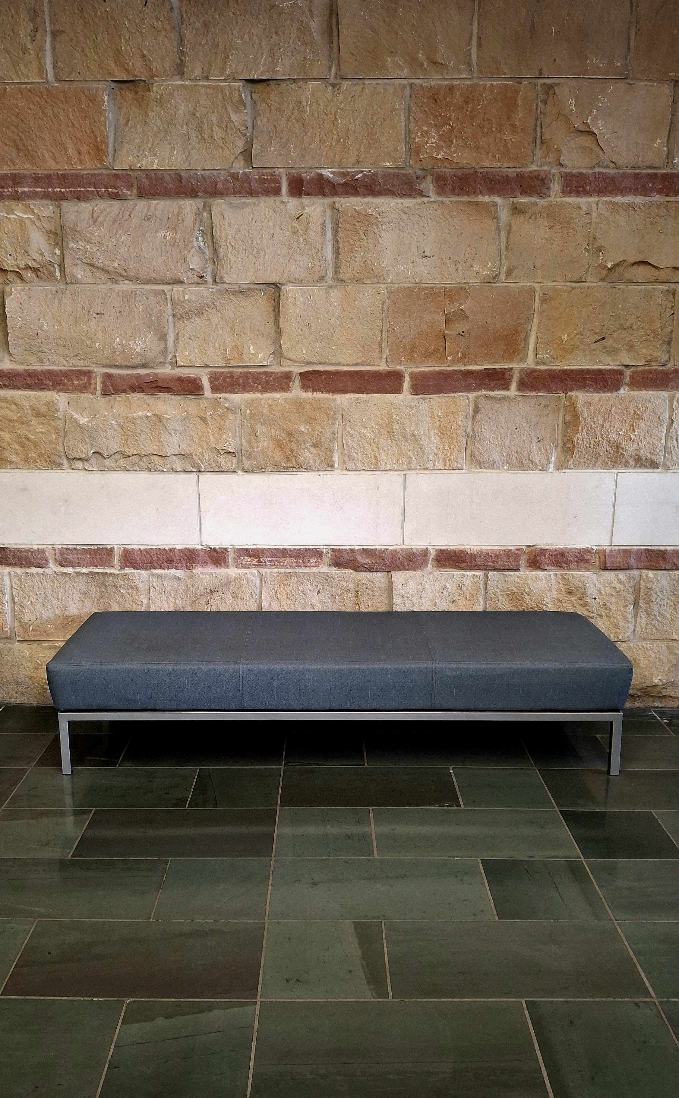 A grey upholstered bench against a wall of layered stone and smooth blocks
