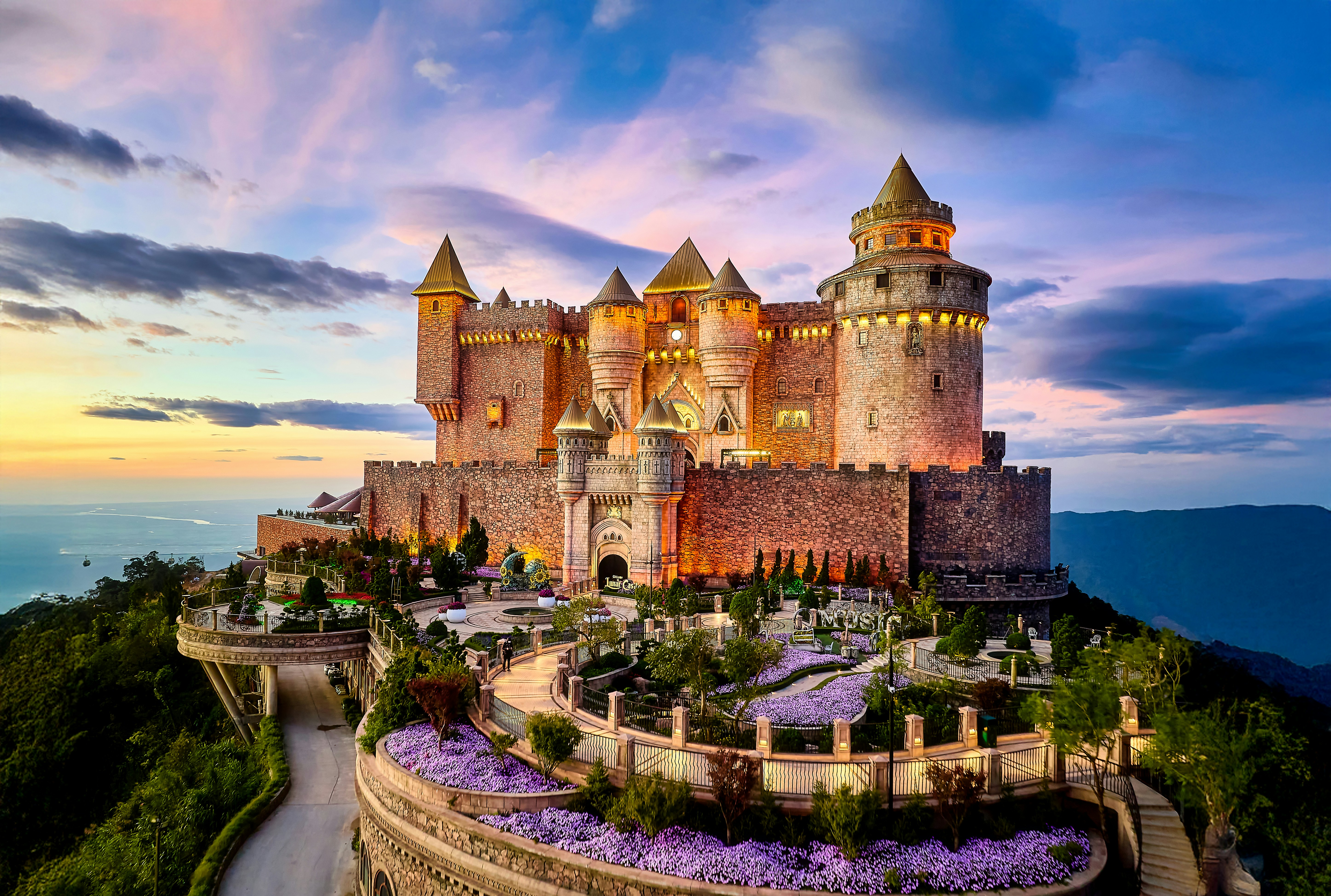 A stone castle on a mountain peak with purple flowers at golden hour