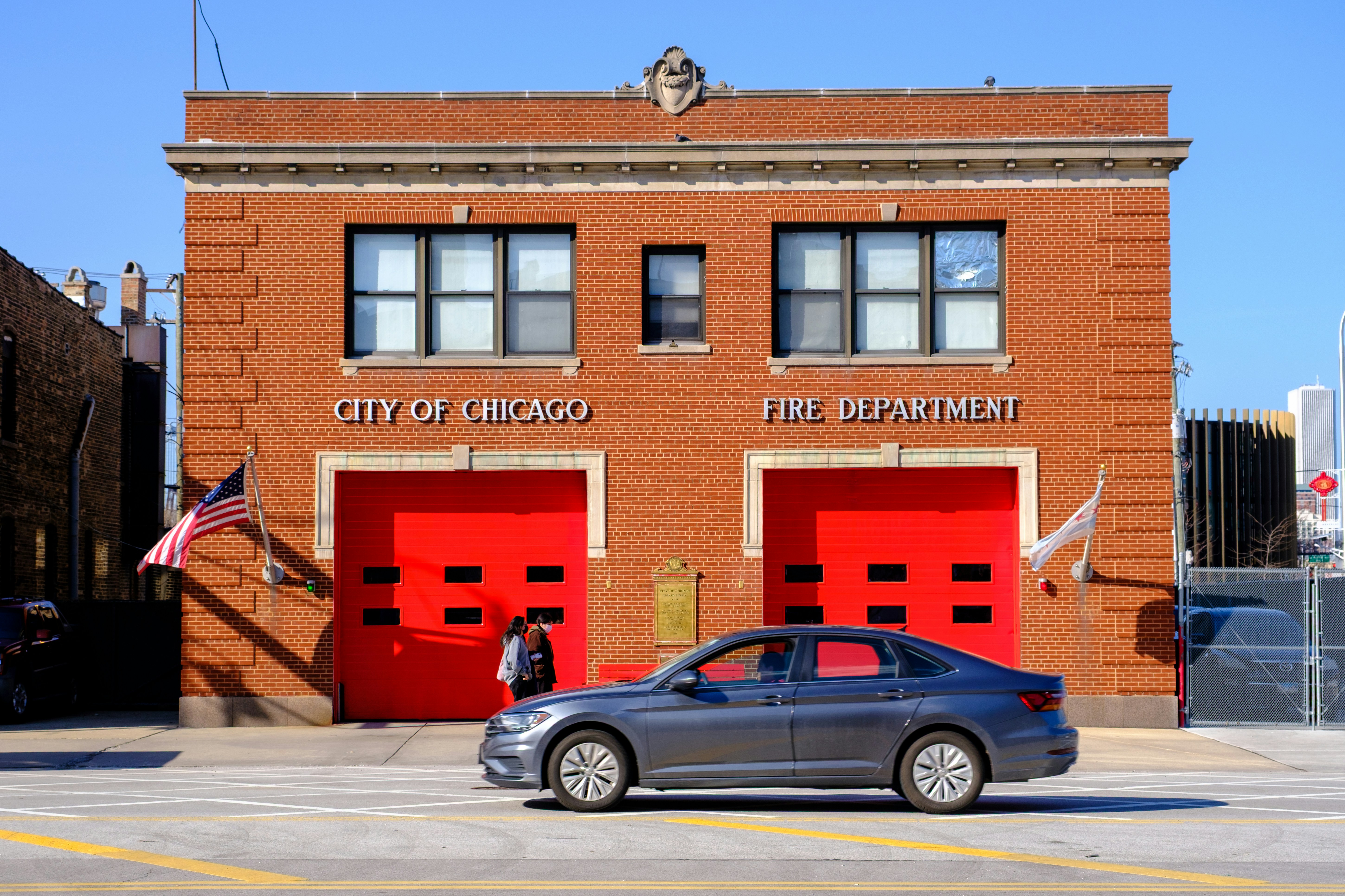 A grey sedan driving past a red brick Chicago fire station with bright red doors