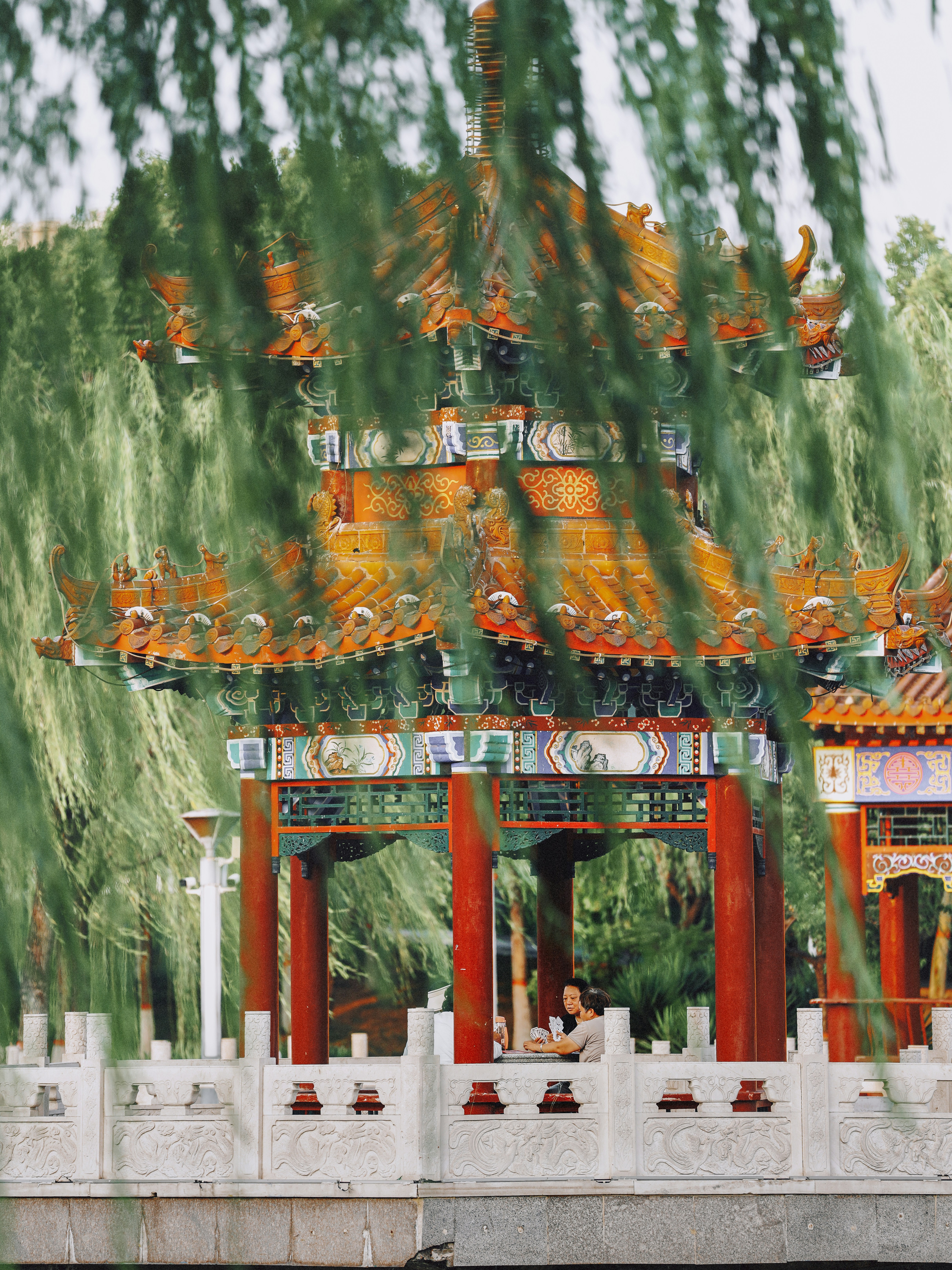 Two people sitting in a traditional Chinese pavilion framed by hanging willow branches