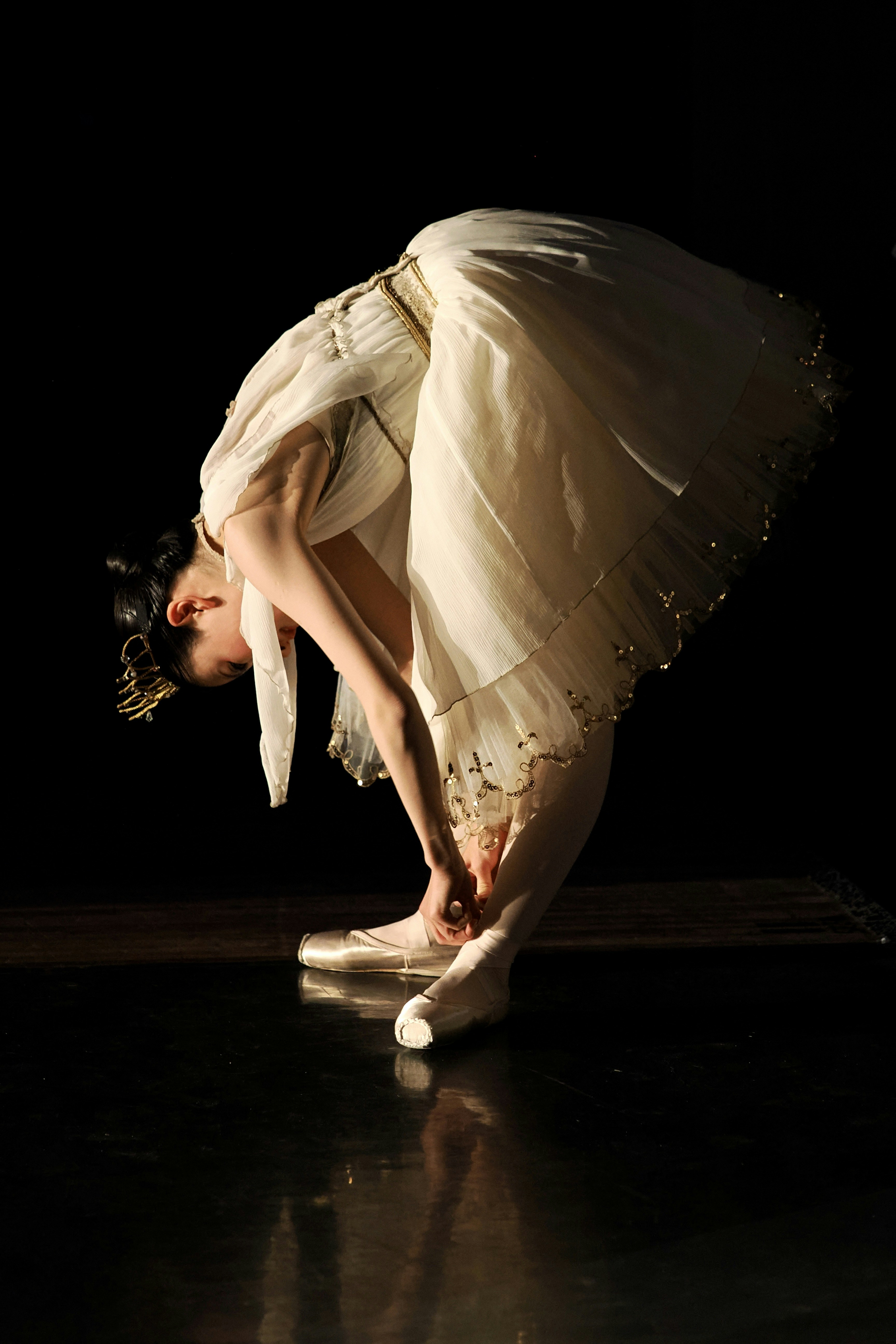 A ballerina in a white tutu bending to tie her pointe shoe