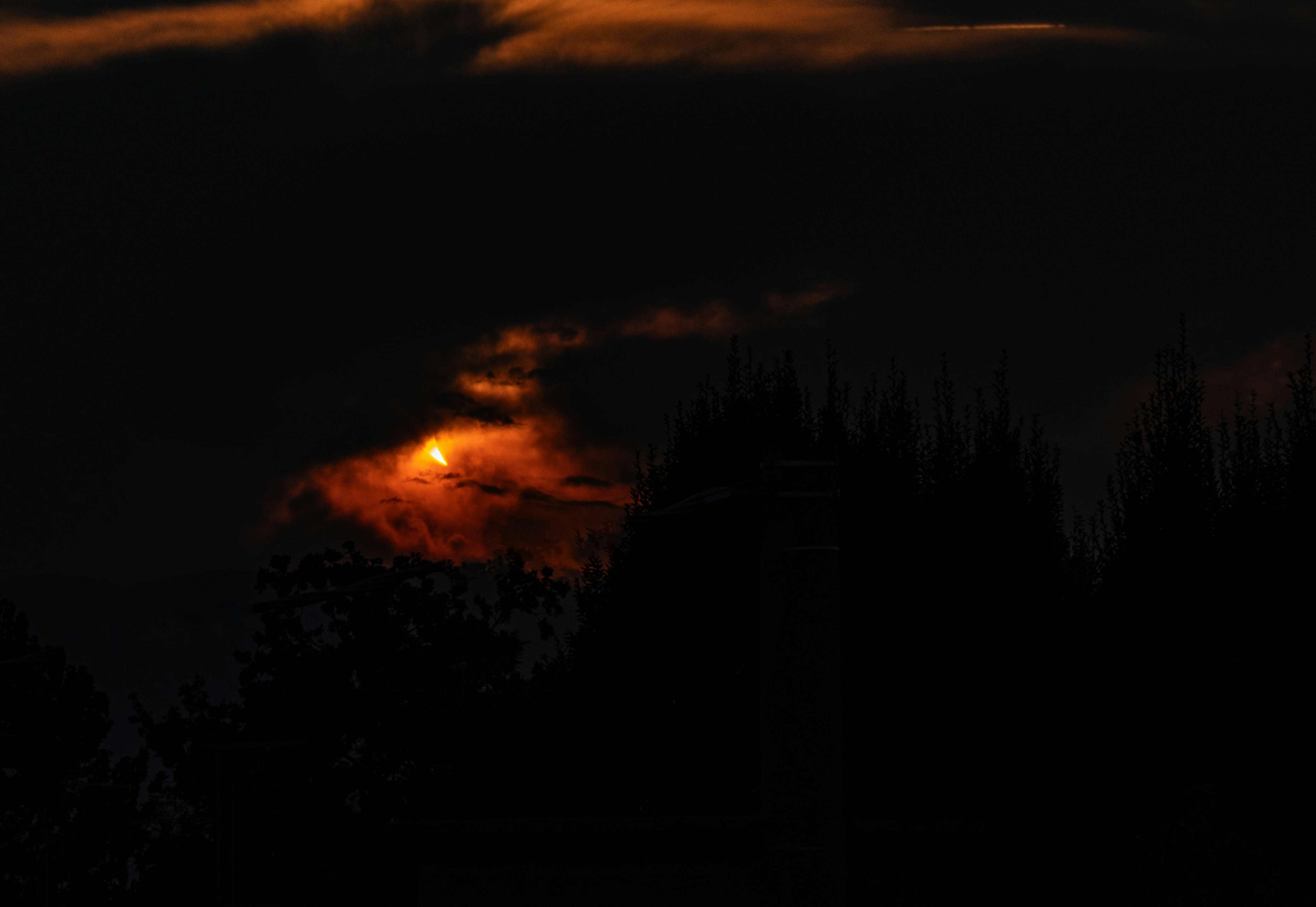 A sliver of light glowing behind dark clouds above silhouetted trees