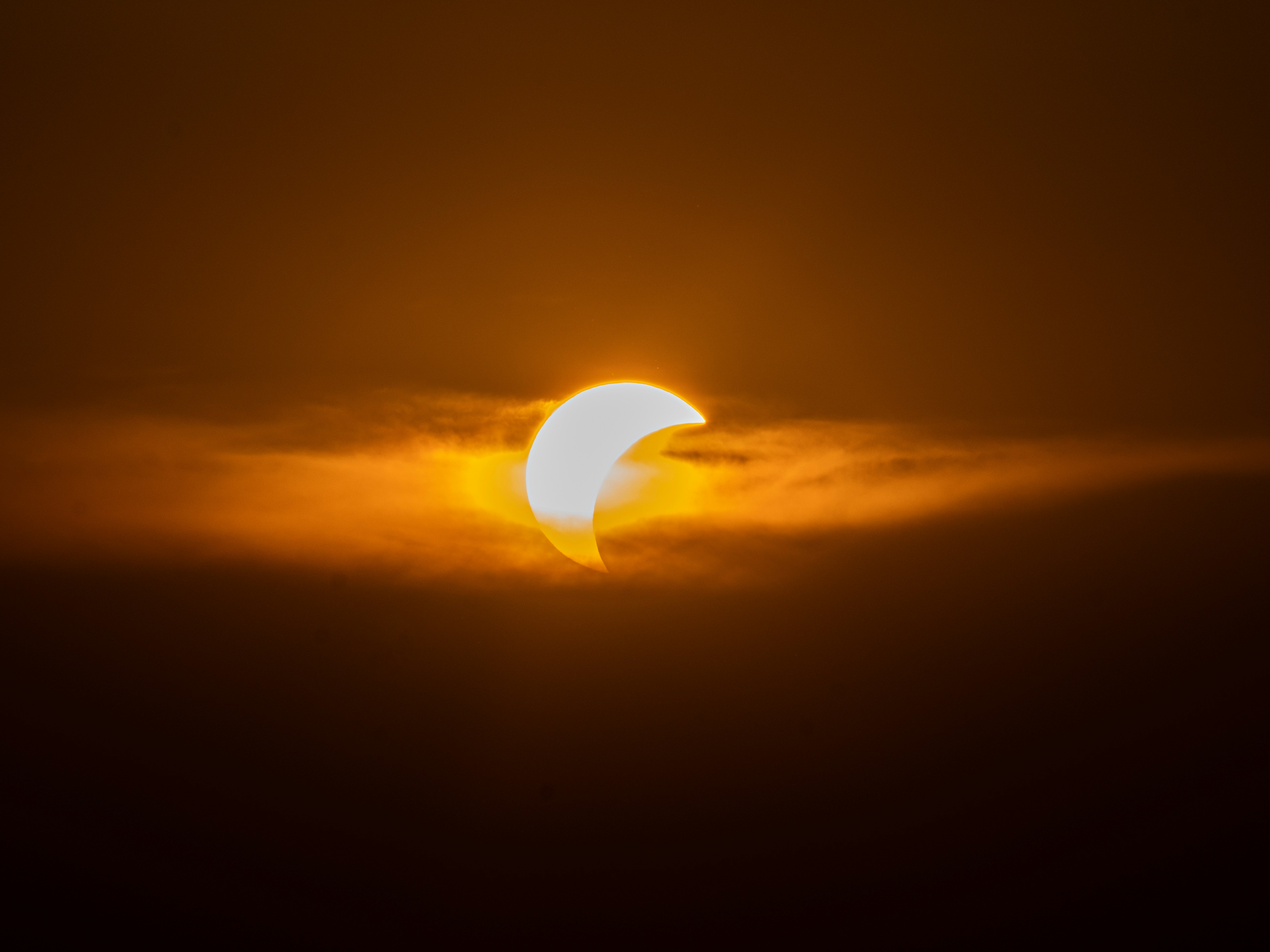 A partial solar eclipse obscured by thin orange clouds in a dark sky