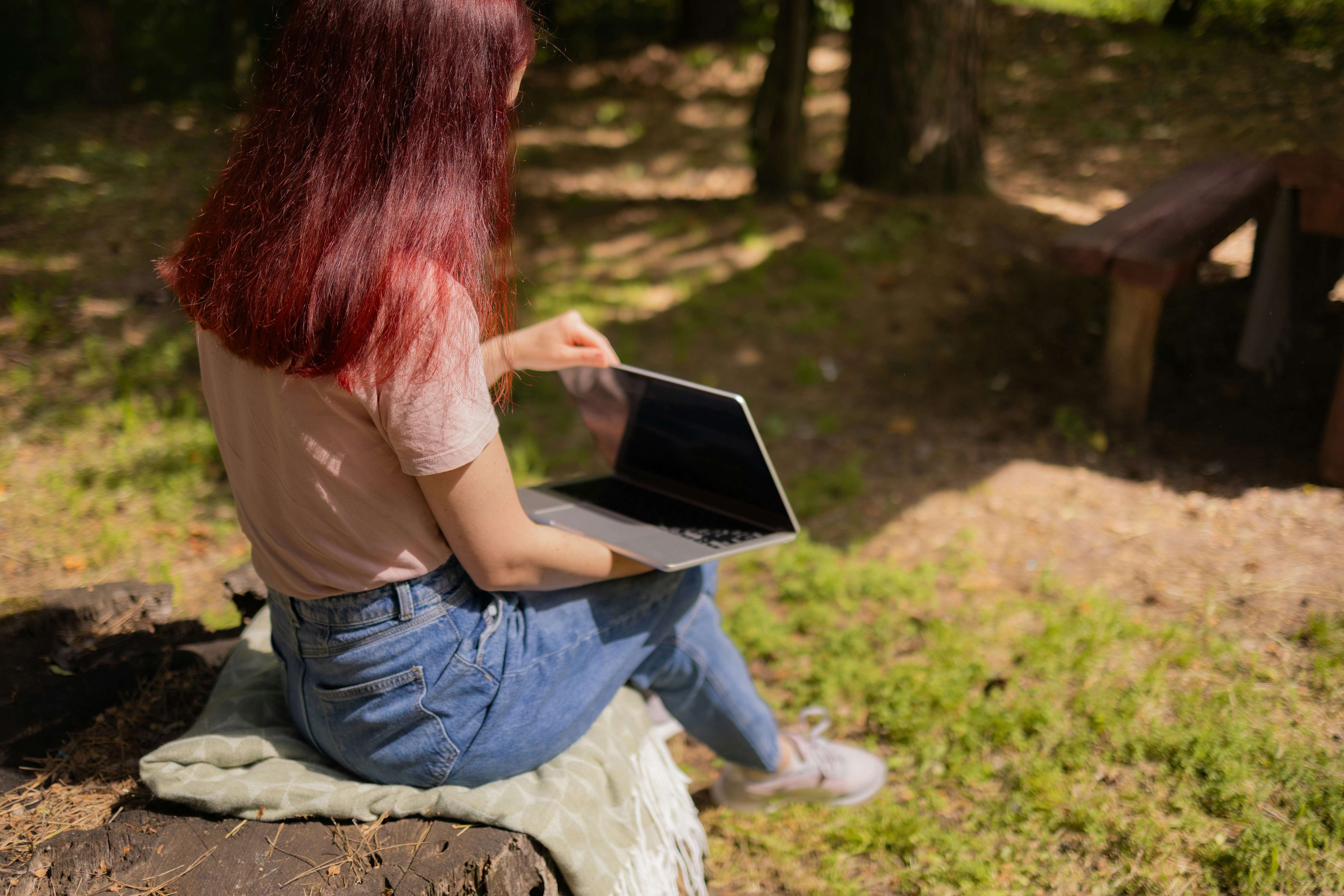 A woman with red hair using a laptop on a tree stump outdoors