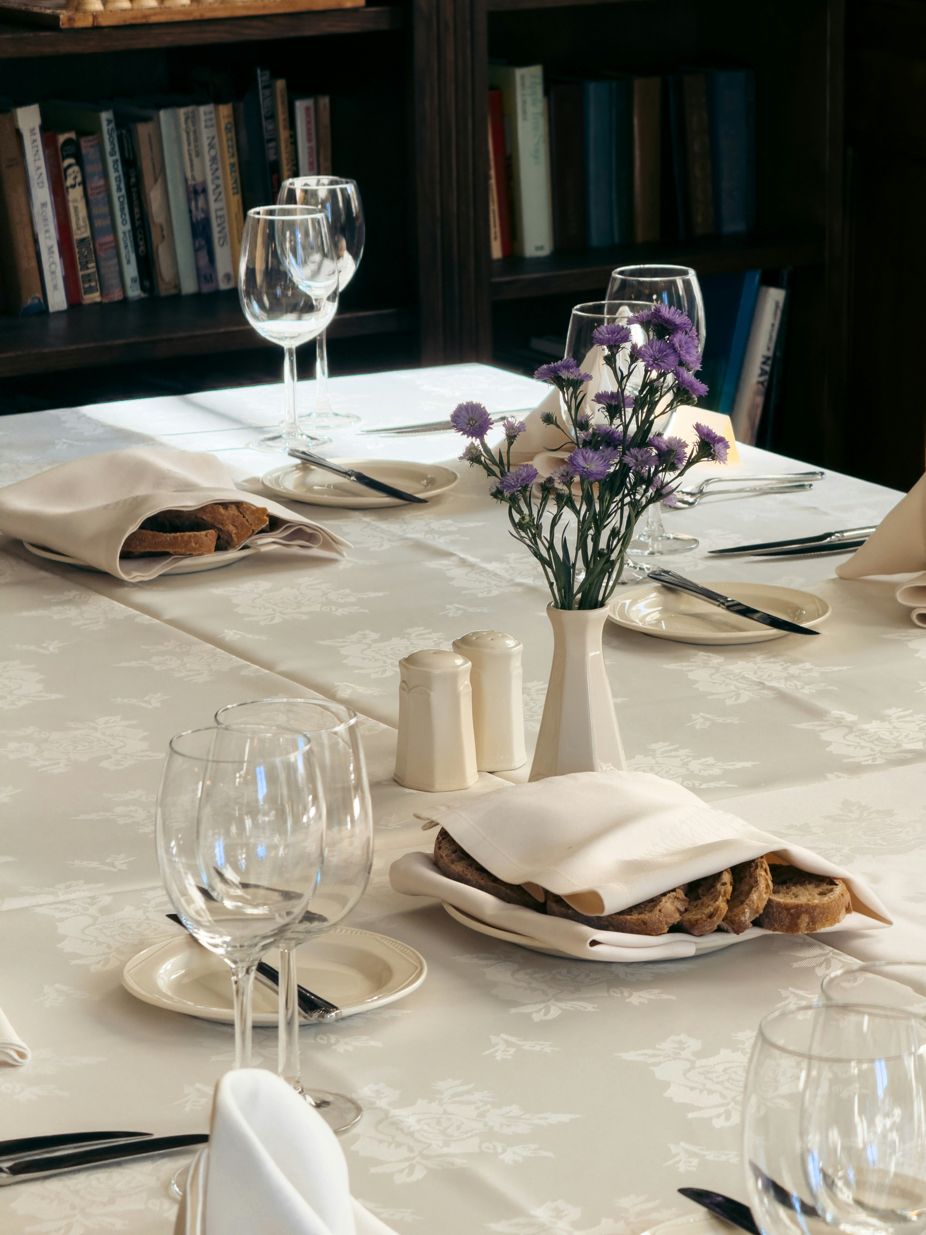 A dining table with bread, wine glasses, and purple flowers in a vase