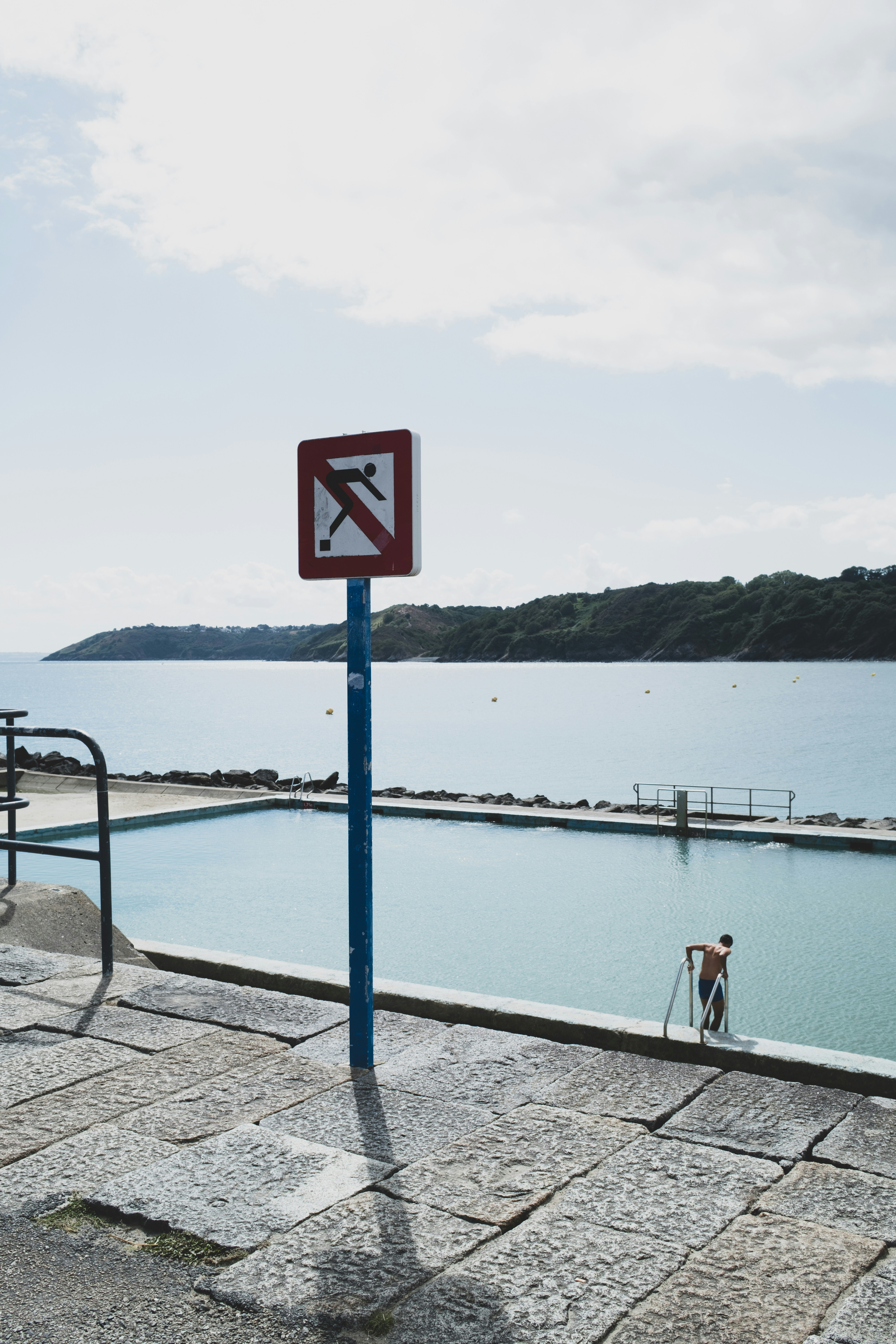 Uma placa de proibido mergulho ao lado de uma piscina externa com uma pessoa entrando na água