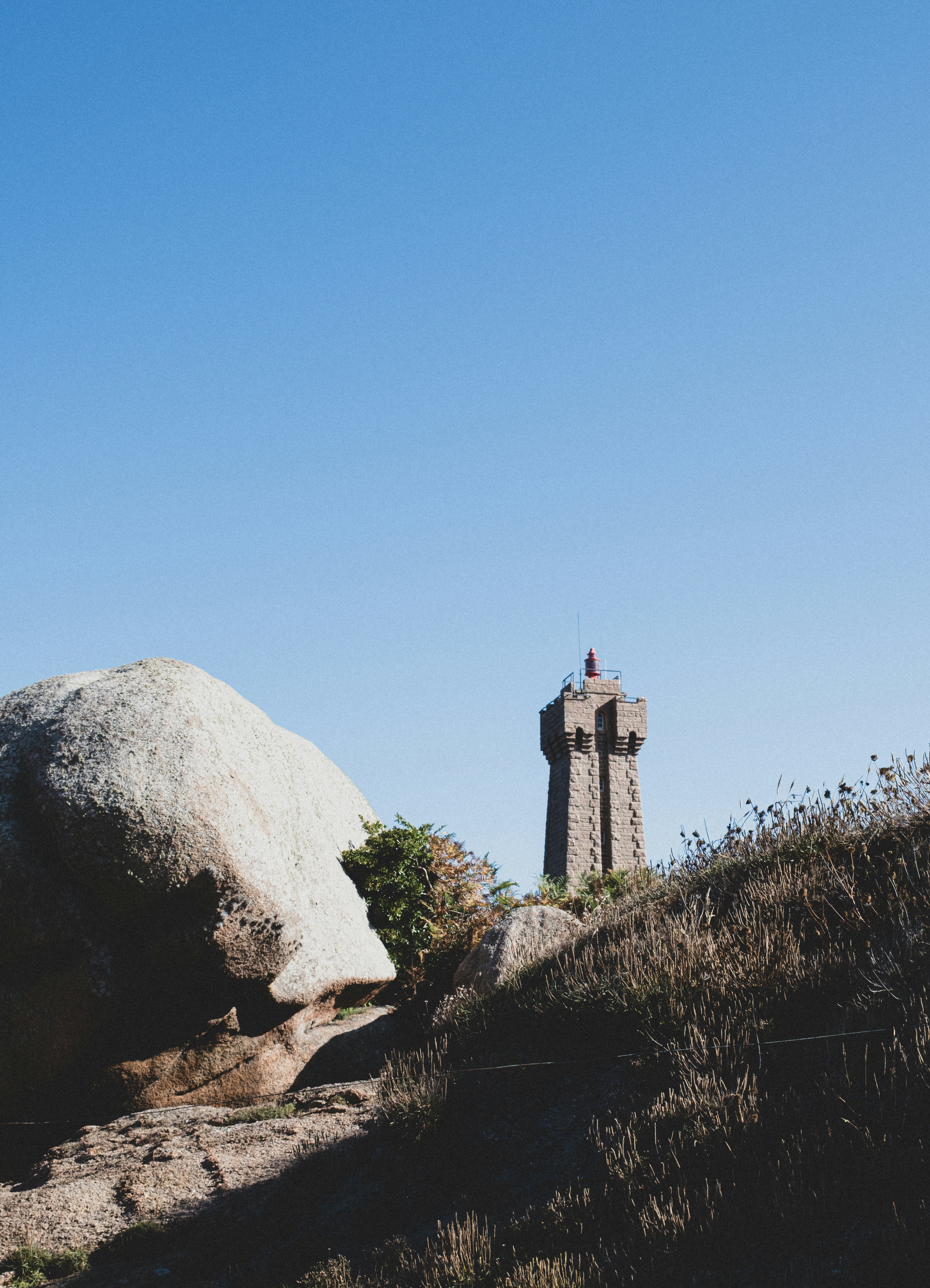 Um farol de pedra atrás de grandes pedras de granito e grama seca sob um céu limpo