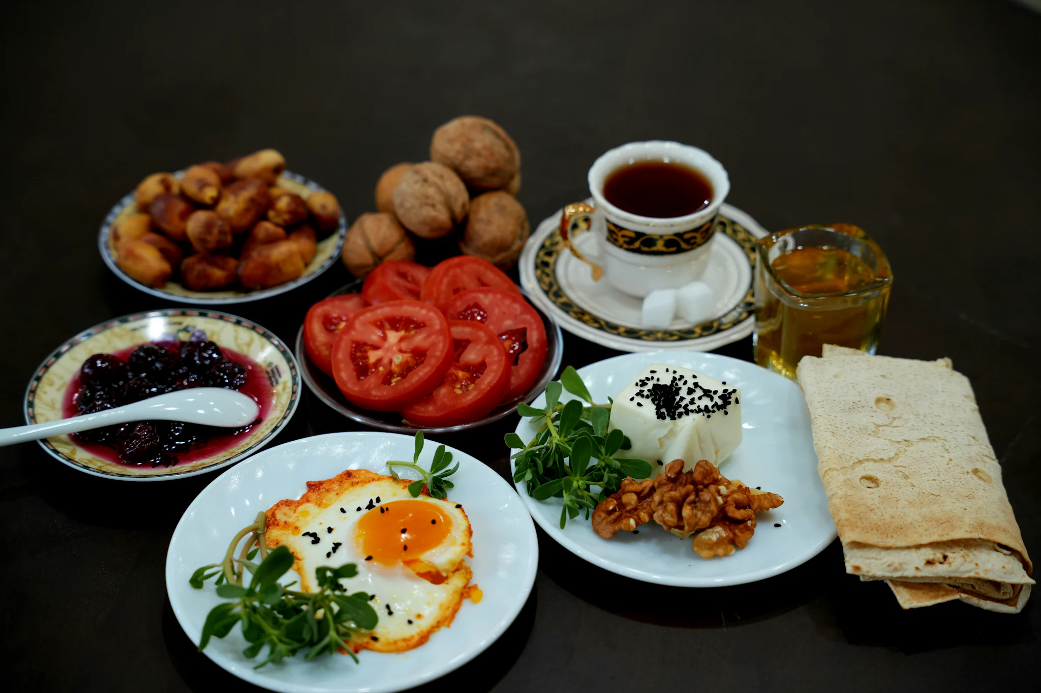 A breakfast spread on a dark table: a fried egg with herbs, sliced tomatoes, white cheese, walnuts, dates, jam, flatbread and a glass of tea