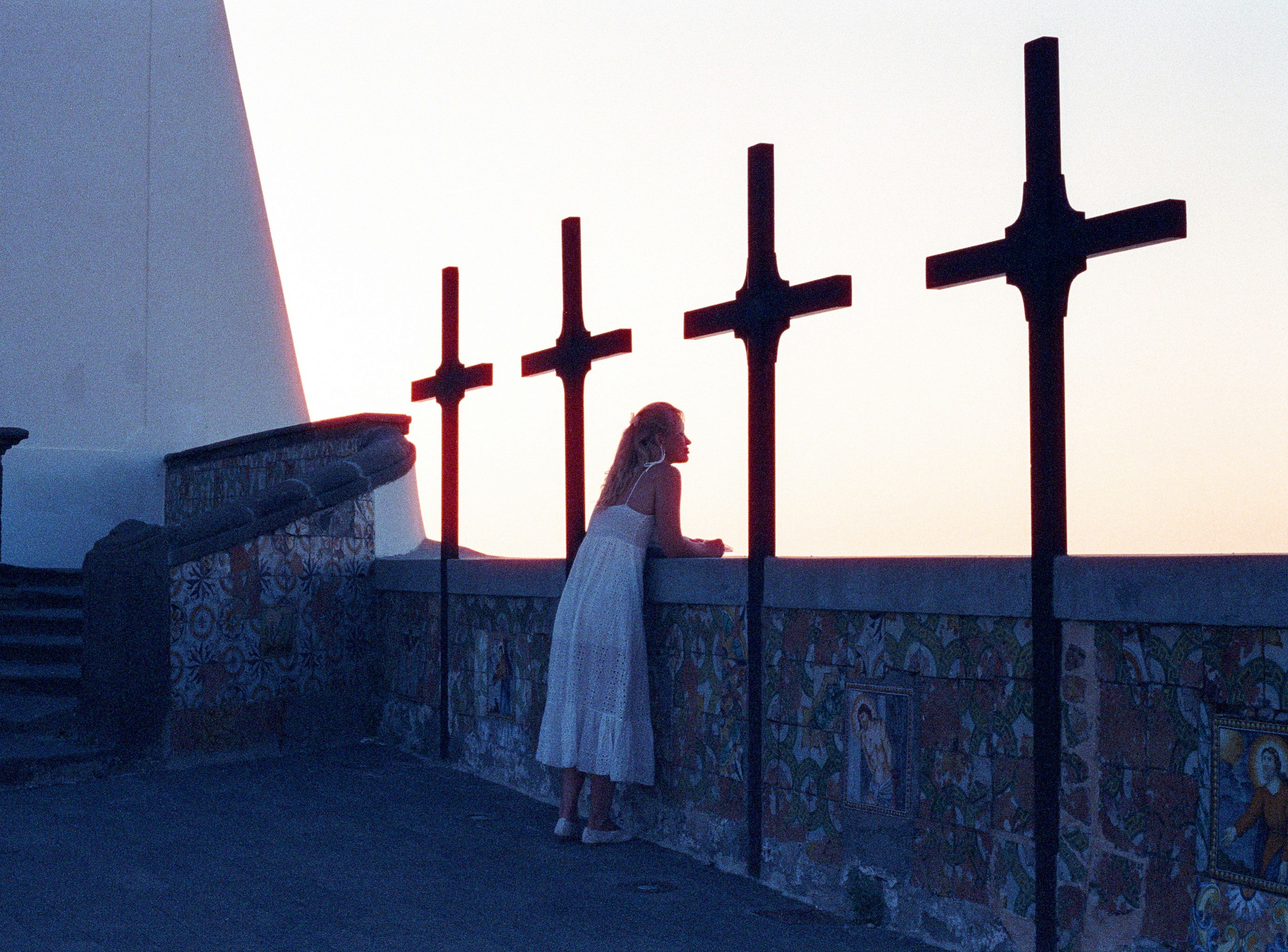 A woman in a white dress leaning against a wall with four crosses