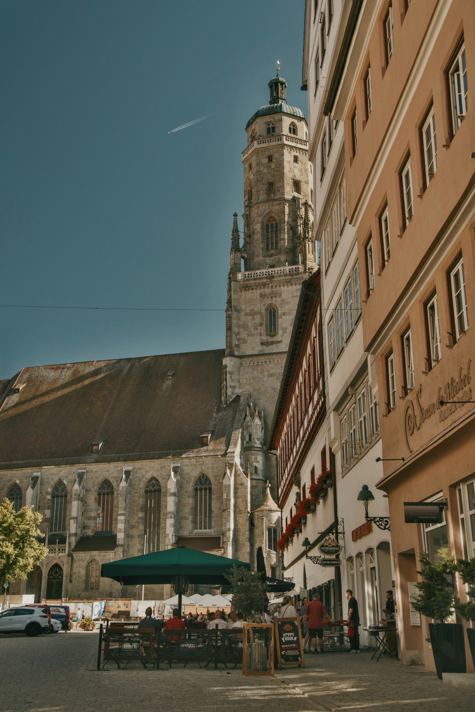 Menschen, die an Außentischen in der Nähe einer hohen Steinkirche in Nördlingen speisen.