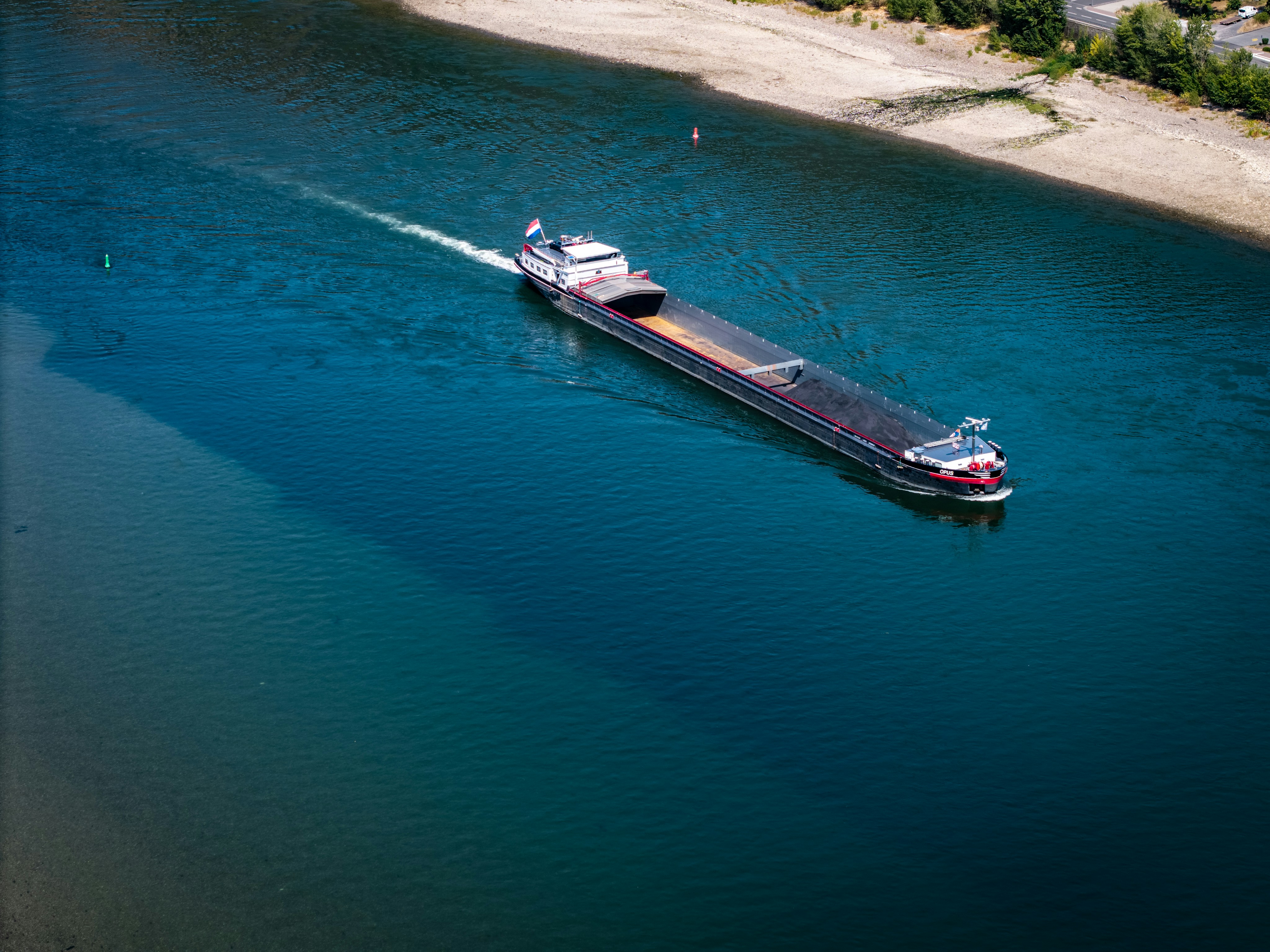 A long cargo barge moving through deep blue river water near a sandy bank