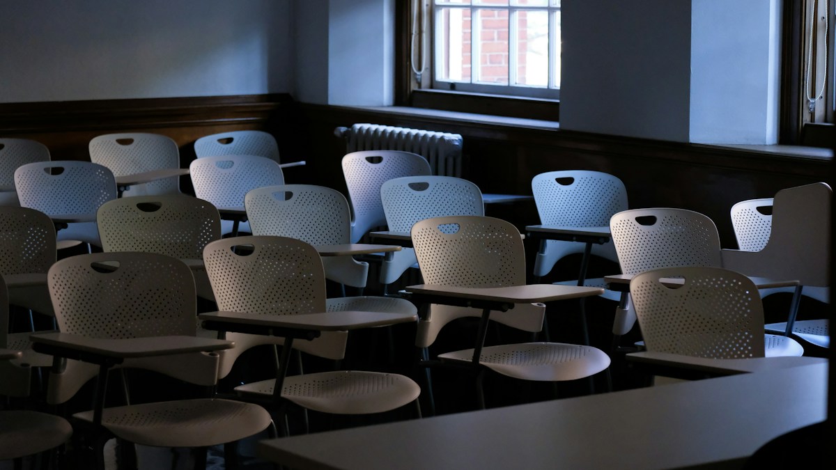 Rangées de chaises blanches à tablette dans une salle de cours vide et sombre, éclairée par une seule fenêtre