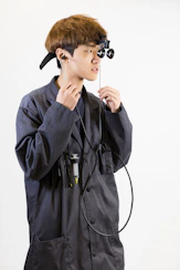 A person in a grey lab coat wearing head-mounted magnifying loupes and holding a stethoscope