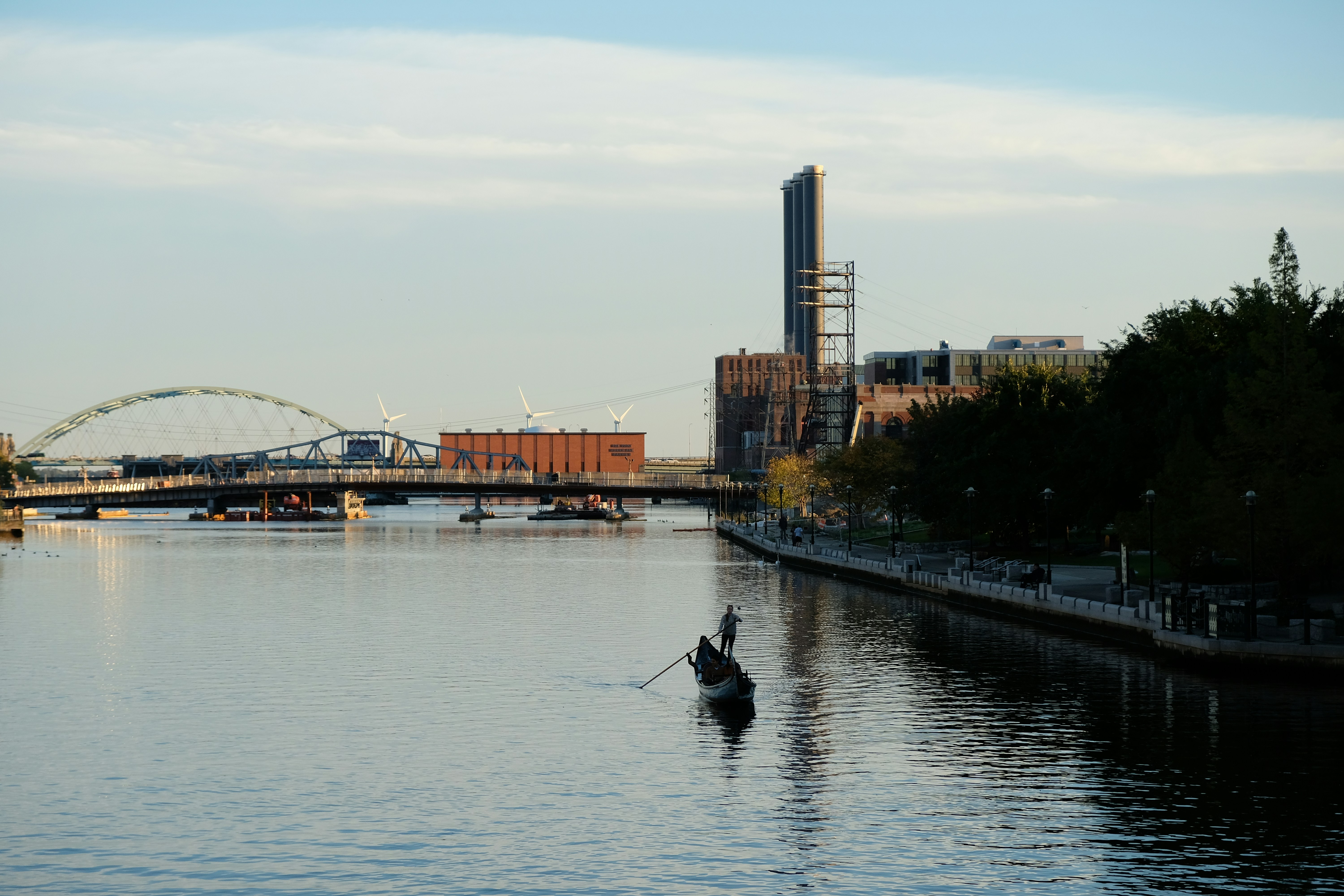 A person rowing a gondola on a river near a bridge and industrial buildings