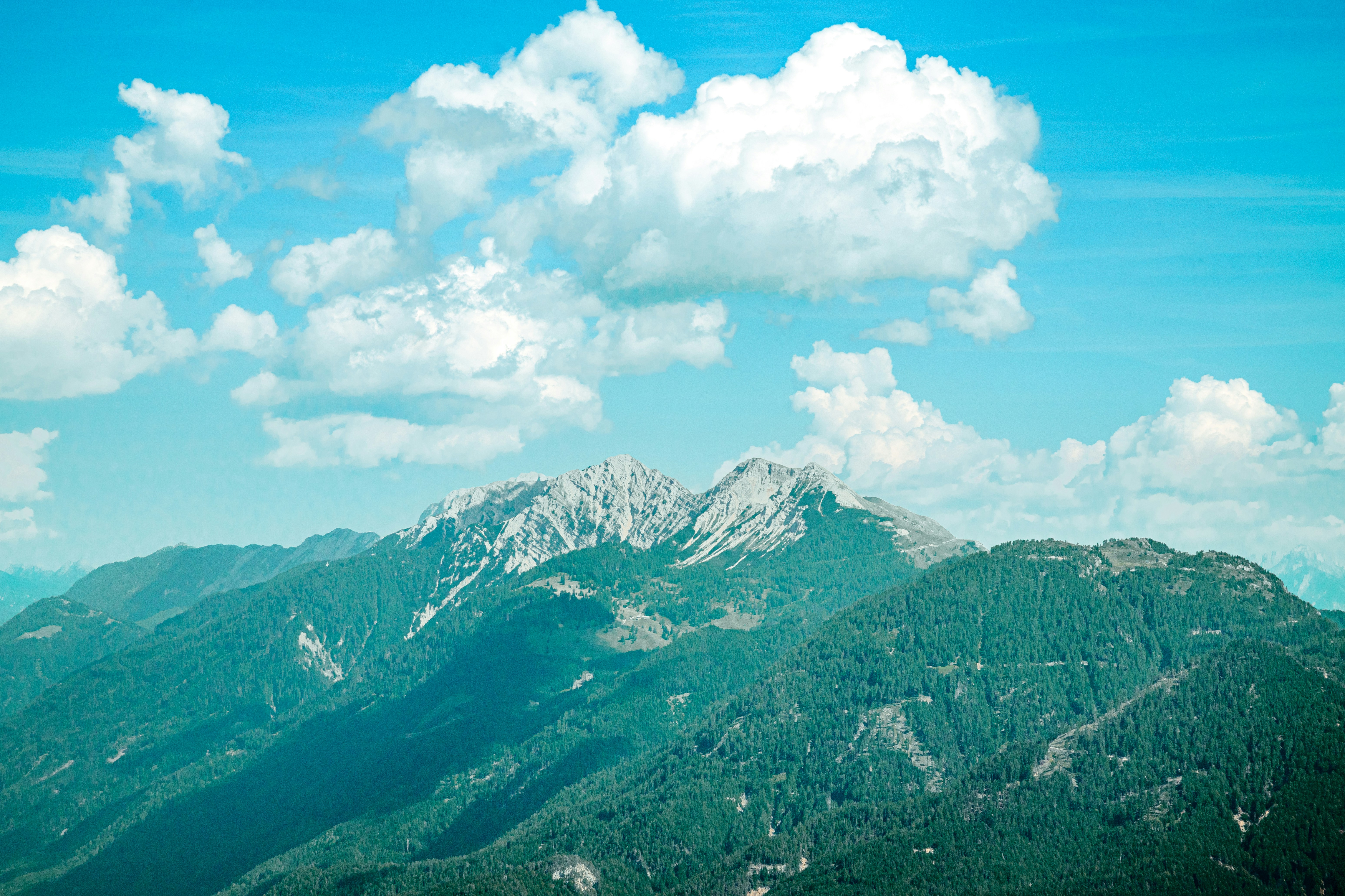 A rocky mountain peak above forested slopes under a bright blue sky with clouds