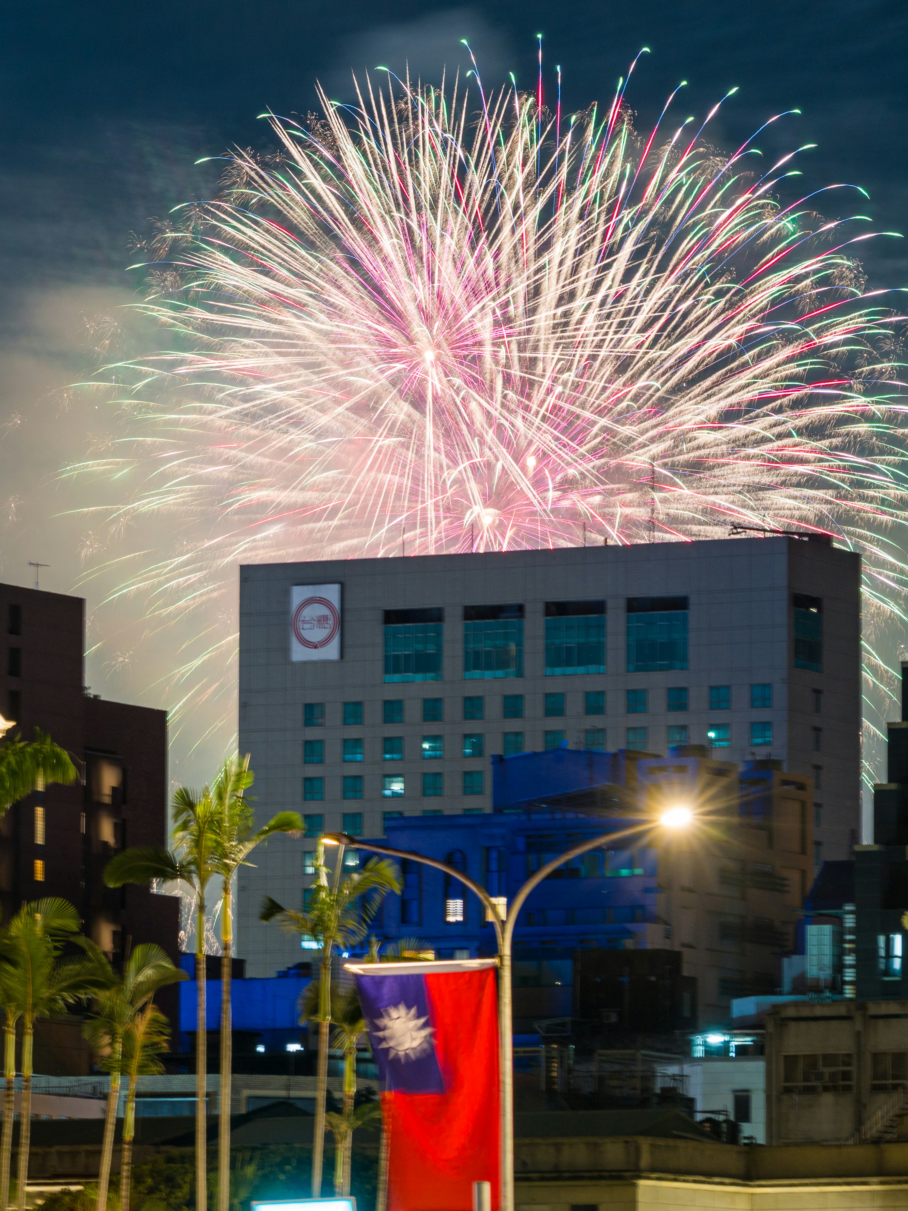 Fireworks exploding over a city building with palm trees and a flag at night