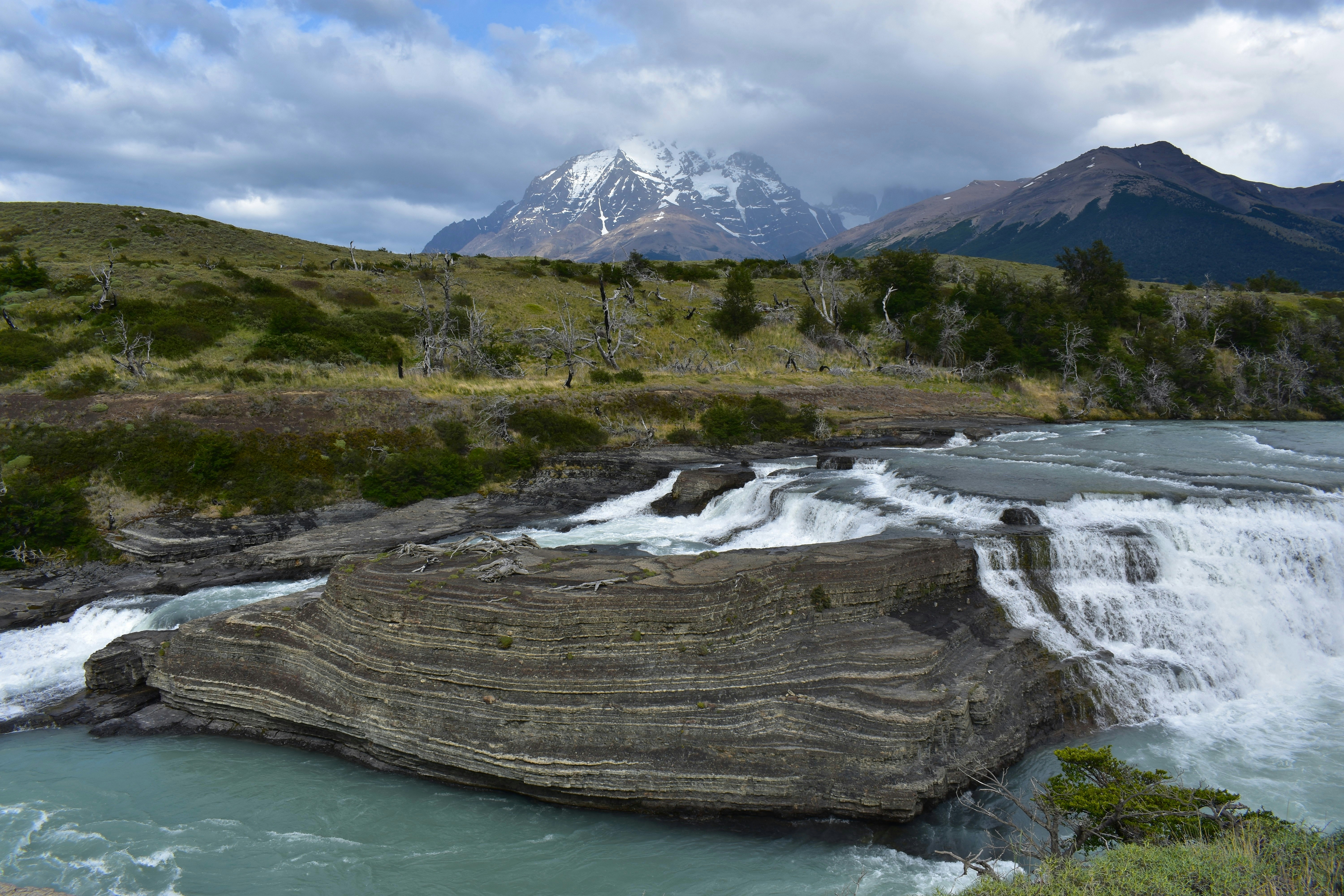 A tiered waterfall flowing over layered rock formations with snow-capped mountains in the distance