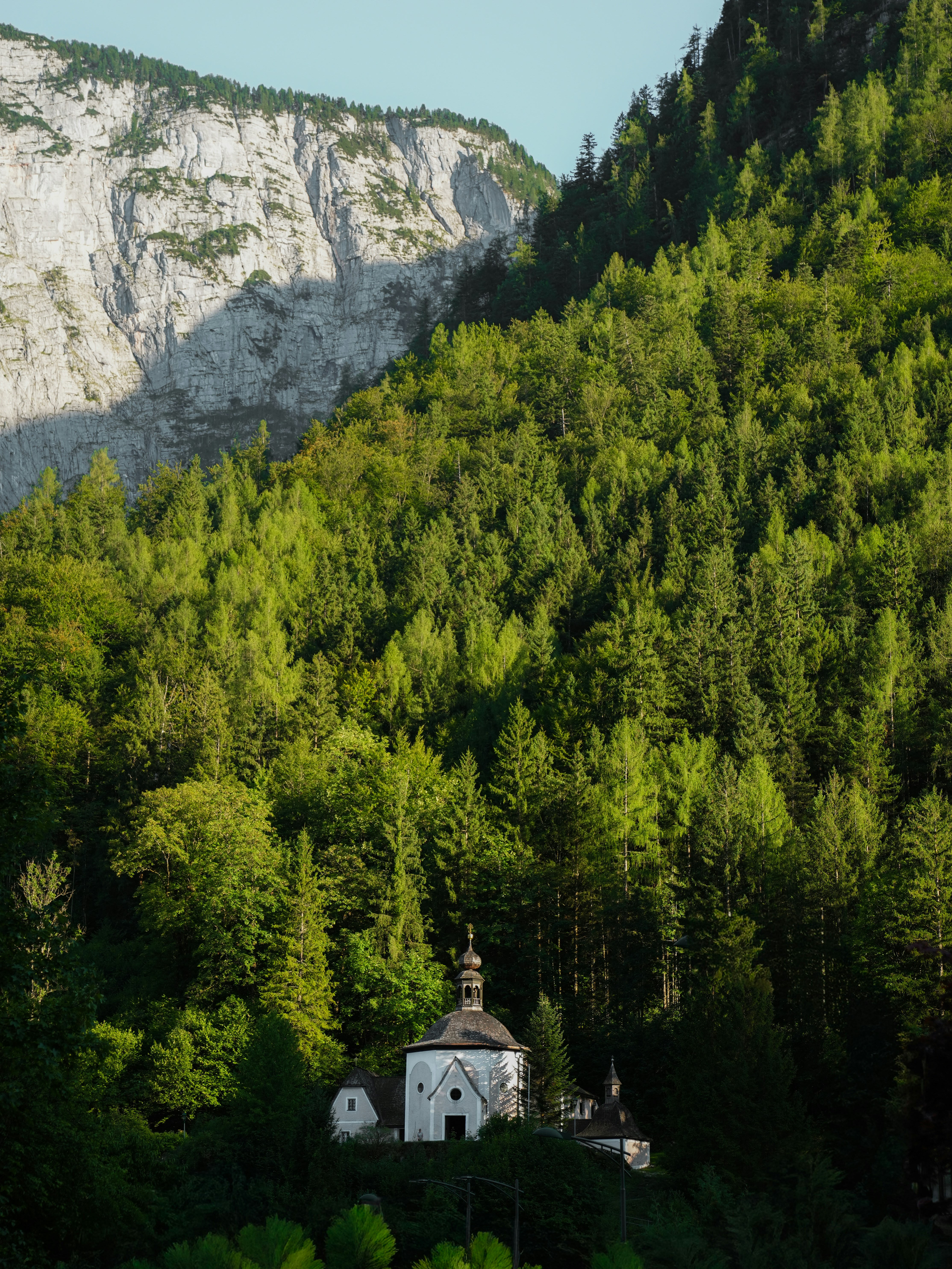 A small white chapel nestled in a dense green forest below a rocky cliff