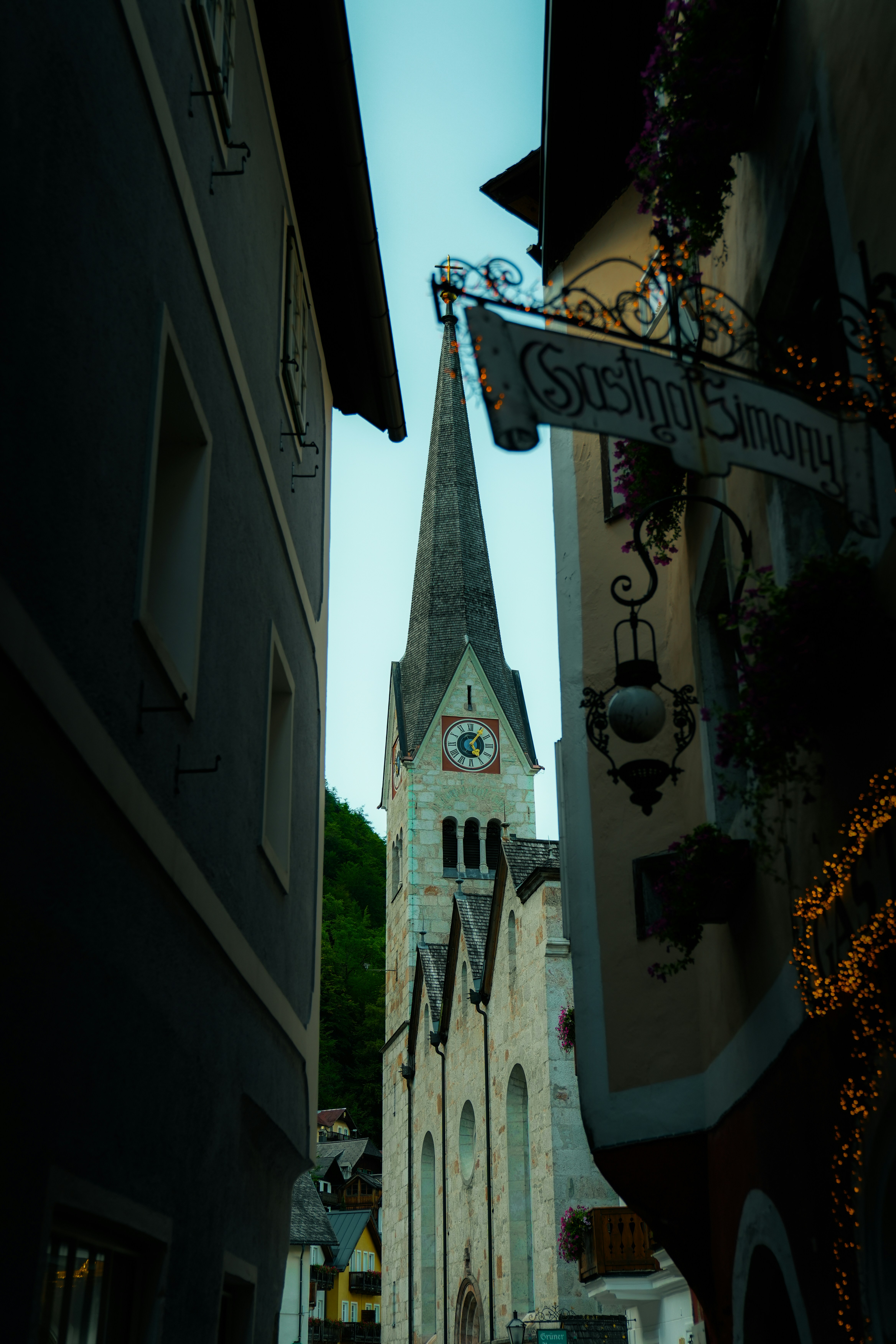 A church spire framed by narrow buildings with a hanging sign in Hallstatt