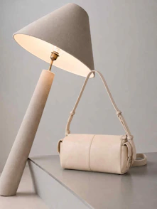 A cream shoulder bag on a grey surface beside a lit floor lamp