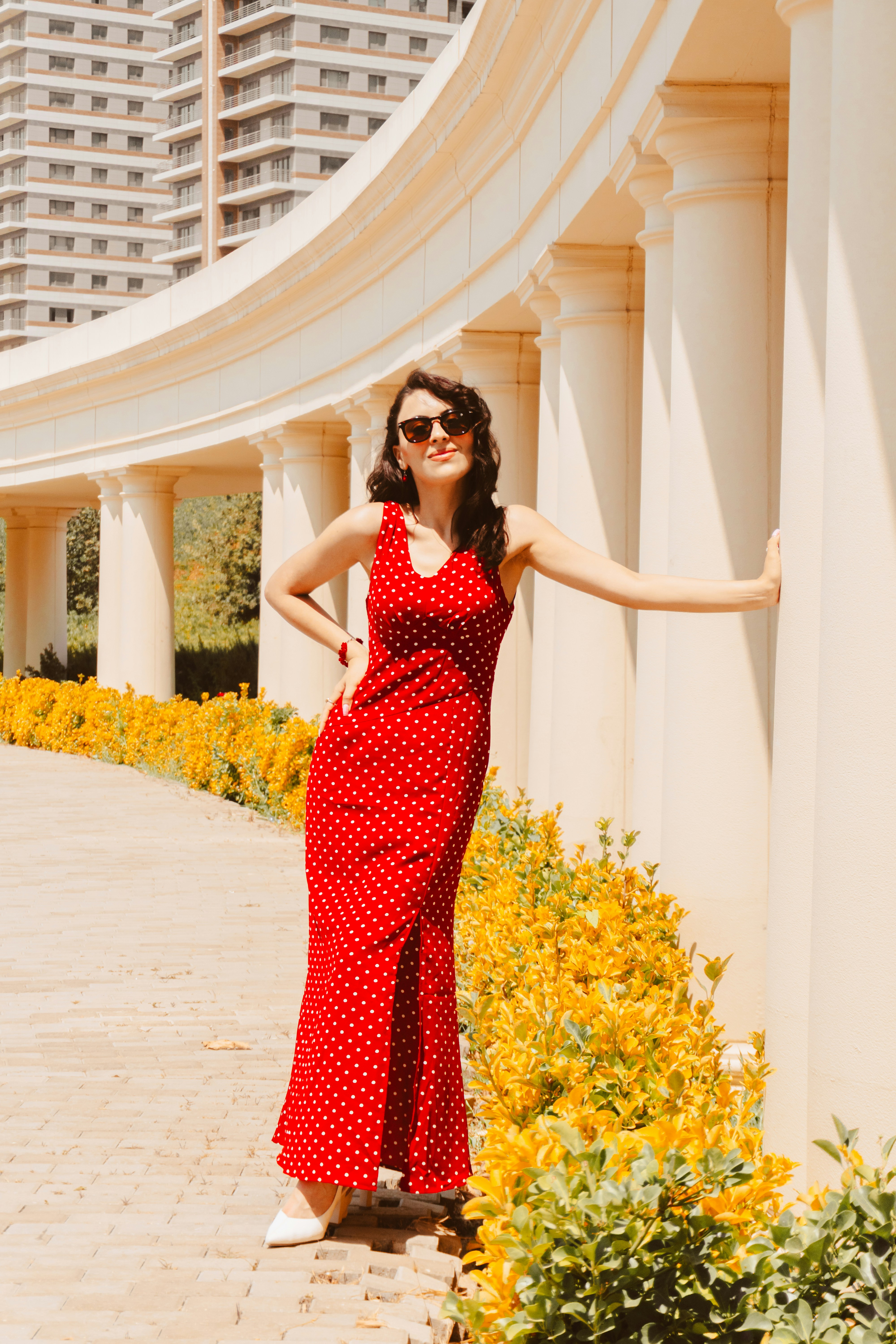 A woman in a red polka dot dress leaning against white columns outdoors