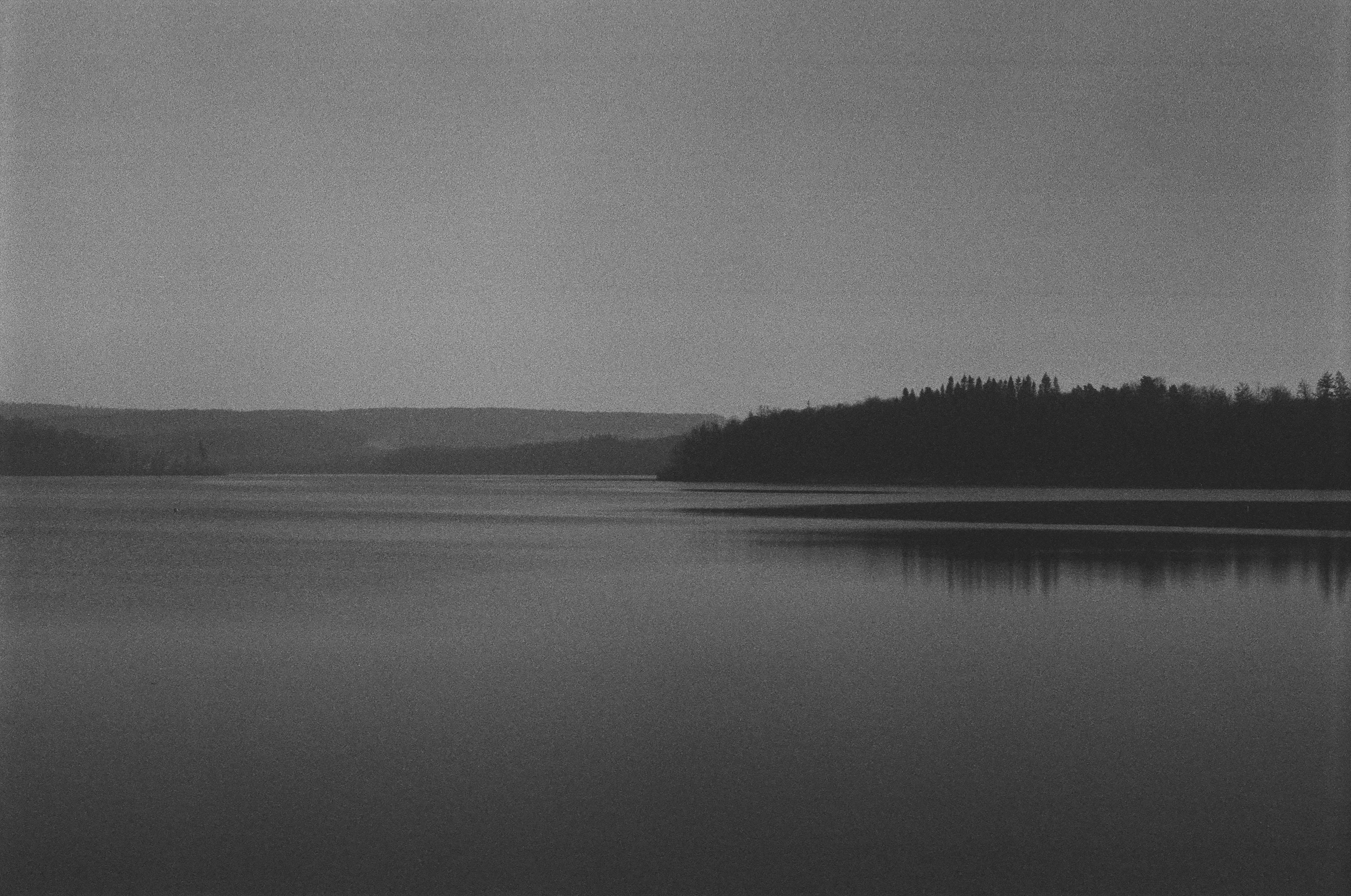 A calm lake reflecting a dark forest under a vast overcast sky