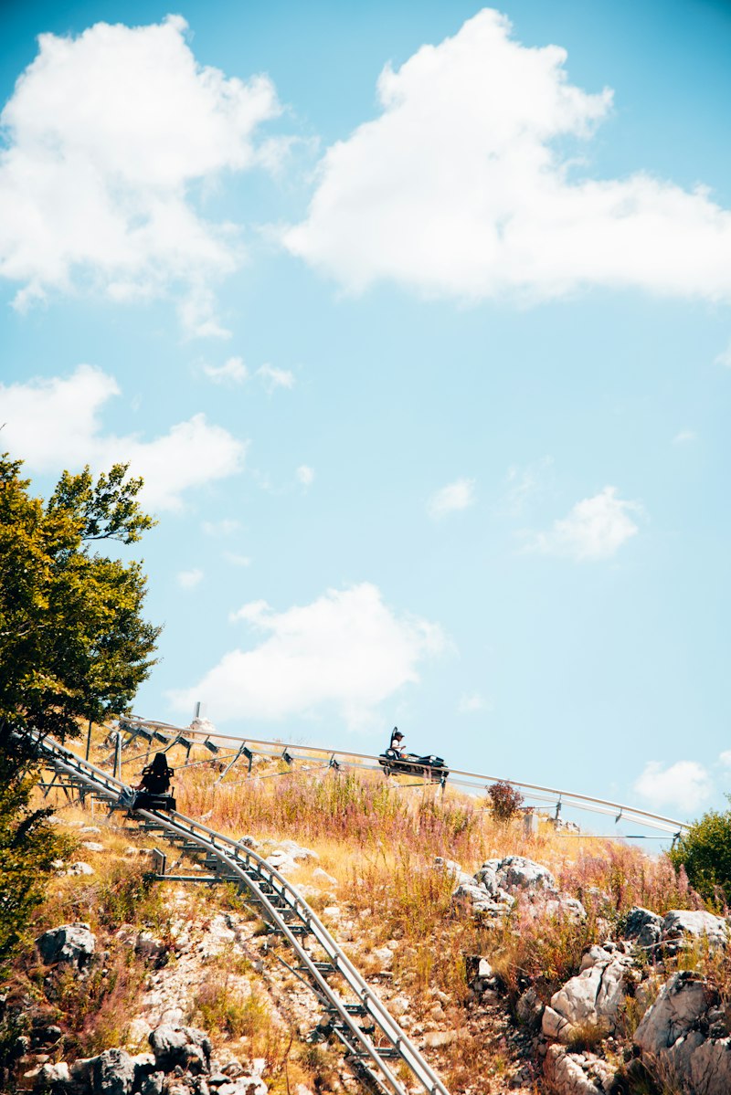 Two riders in a small coaster cart on an elevated steel rail curving across a green mountainside