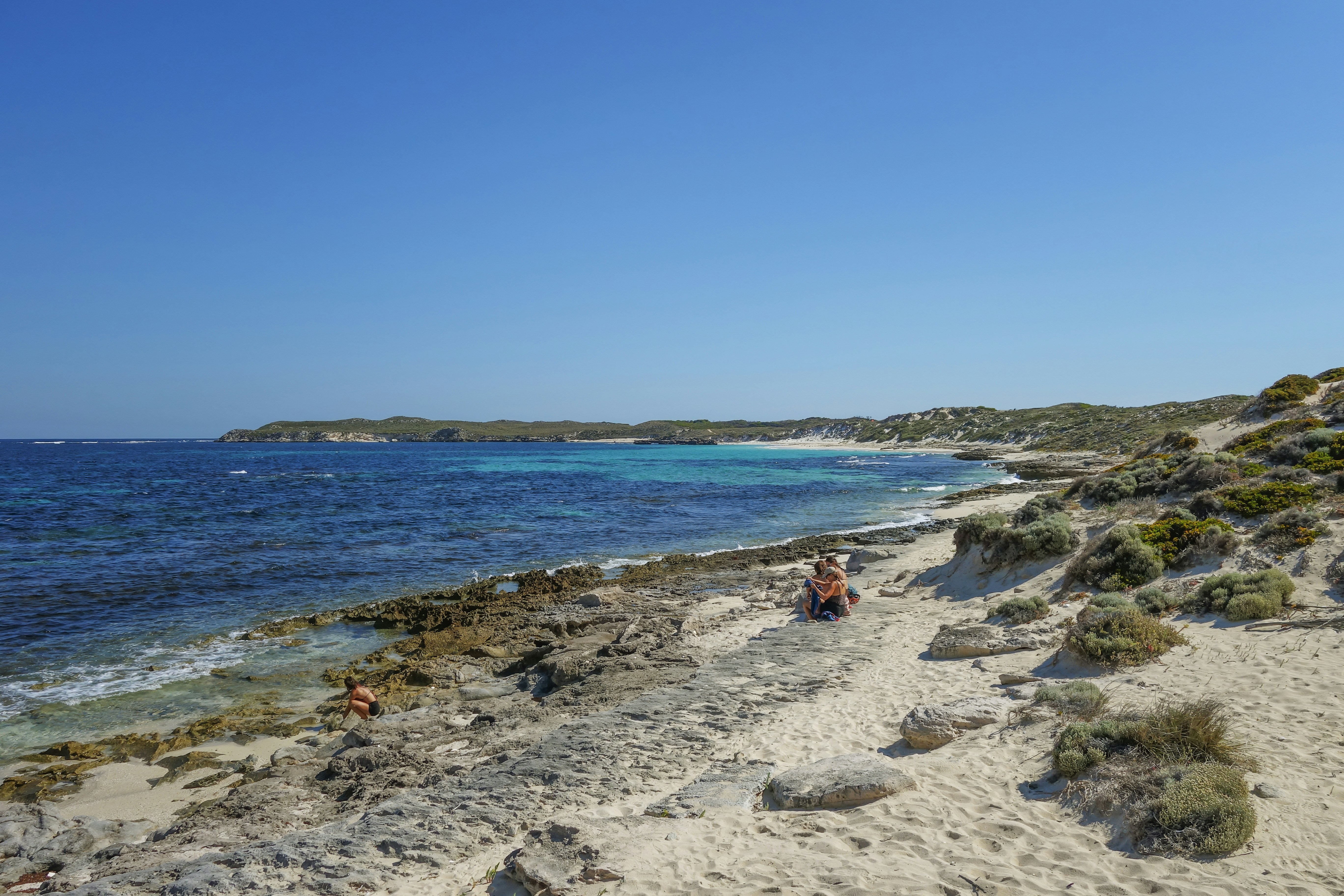 People on a rocky beach with turquoise water and coastal dunes under clear sky